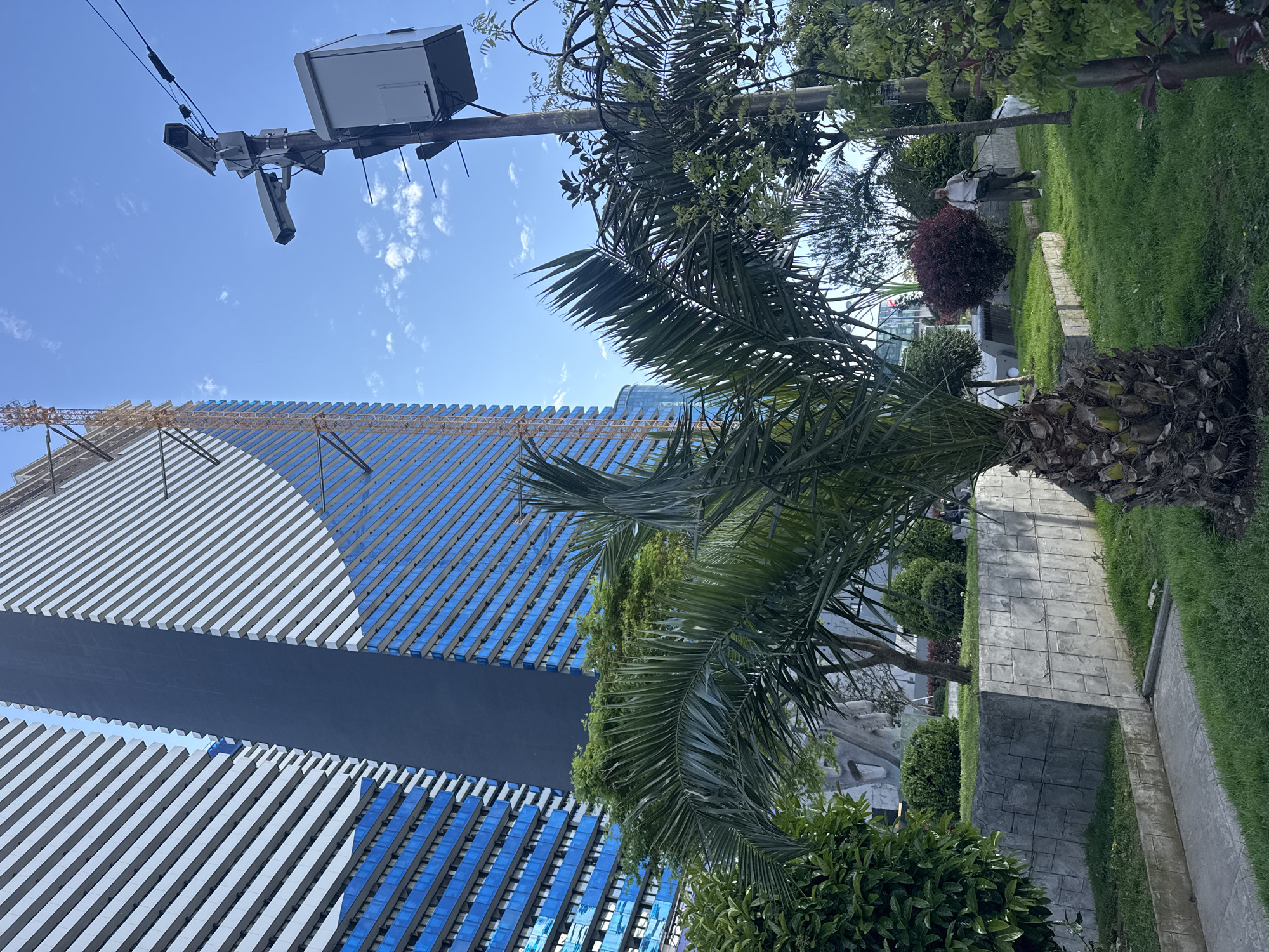 The image captures an outdoor scene on a clear, sunny day, featuring a modern building, lush greenery, and a utility pole, viewed from a slightly low angle.
Dominating the left side of the frame is a contemporary building with a striking facade. It features wide horizontal bands of white and reflective blue, separated by a dark blue horizontal strip running across the middle. A large construction crane is visible above the building, extending towards the top left.
In the foreground and midground, several palm trees with prominent fronds partially obscure the building and sky. Other green bushes and trees are also present, including a small reddish-purple bush in the midground. To the right, a well-maintained grassy area borders a patterned stone-like concrete pathway or low wall. A single person, dressed in light clothing, is visible in the midground on the right, walking on the grass.
High above, against the bright blue sky with scattered wispy white clouds, a utility pole is equipped with various devices, including a large white box and multiple dark, rectangular sensors or cameras.
The scene depicts a calm, everyday moment in what appears to be an urban or park-like environment, suggesting ongoing development with the visible construction crane. No discernible text is present.