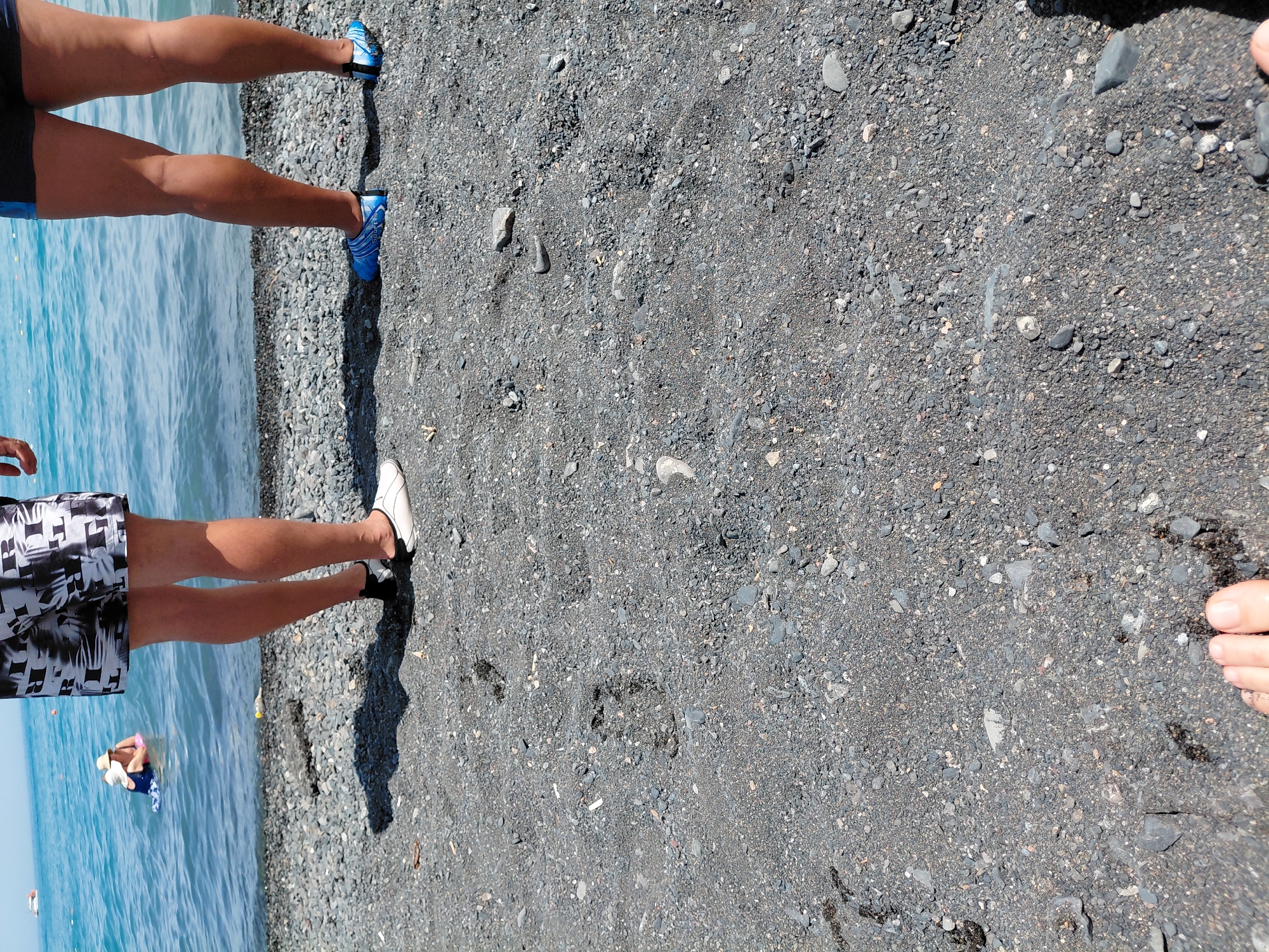 The image depicts a beach scene with dark sand and pebbles. In the background, the blue water of a sea or large lake is visible. Two individuals are partially visible standing on the shore, facing away from the camera. The person on the left wears black and white patterned swim shorts and white water shoes. The person on the right wears dark swim shorts and blue water shoes. In the water, a third person, possibly a child wearing a blue swimsuit and a straw hat, is floating on a pink inflatable ring. The environment appears to be a sunny day. The location is specified as Ақши, Kazakhstan, suggesting this is a beach on Lake Balkhash.