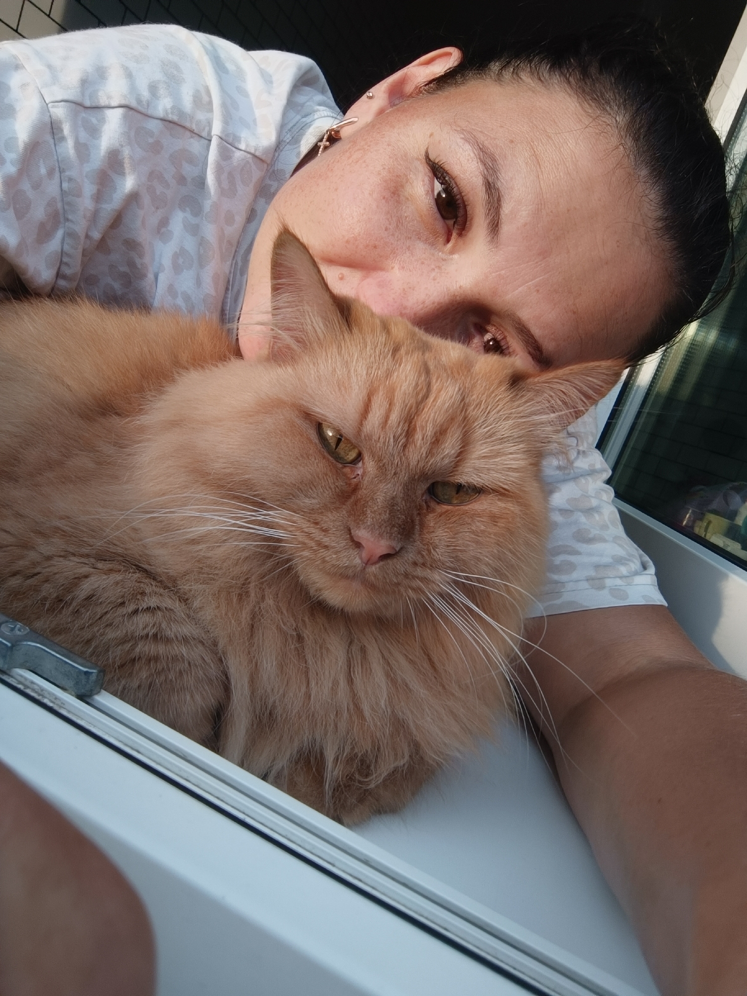 A close-up selfie shows a woman with dark hair and light brown eyes leaning her head against a fluffy orange cat. The woman is wearing a white shirt with a light grey leopard print. The cat is looking forward with its eyes partially closed. They are positioned on a white windowsill, with a window frame visible in the background. The scene appears to be indoors, likely in an apartment in Chaikovsky, Russia, as indicated by the context. The lighting suggests it is daytime, with sunlight casting shadows.