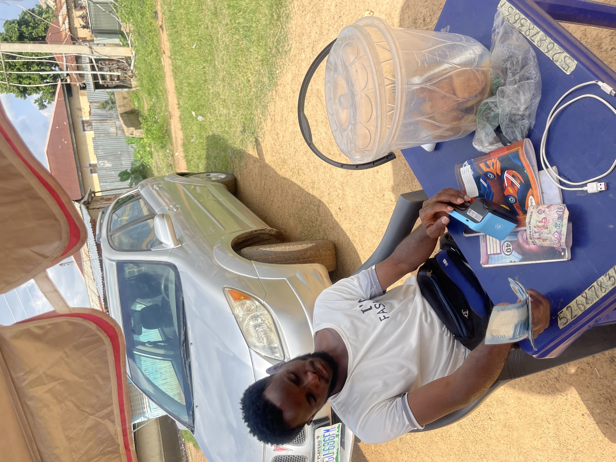 A young Black man with a beard and short, curly hair is seated in a dark chair next to a blue plastic table. He is wearing a white t-shirt with "I'M FASHION" printed on it in black, and a blue and black fanny pack around his waist. His eyes are closed, and his head is tilted down as if he is resting. The man holds a small stack of Nigerian Naira bills in his right hand, with one bill clearly visible showing "1000" and text from the Bank of Nigeria.
On the blue table are several items: a notebook with a design of two race cars, a blue card reader, a plastic bag containing some food items, a clear plastic bucket with a lid containing more food items and a spoon, and a white USB cable.
Behind the man is a silver Pontiac Vibe car with a license plate reading "PLATEAU GU 686FM". The car is parked on a dirt path. In the background, there are several buildings with corrugated metal roofs and fences, as well as lush green grass and trees. The scene appears to be outdoors, under a shade provided by a beige and red awning. The location is identified as Rafin Par, Nigeria. The overall lighting suggests it is daytime.