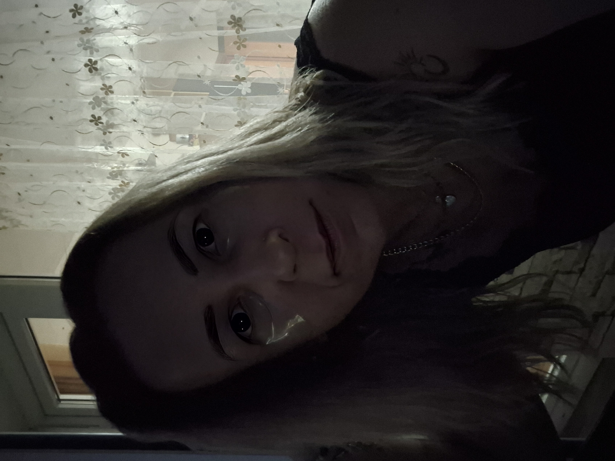 A woman with blonde hair is looking at the camera in a dimly lit indoor setting. She is wearing a dark top and a necklace. There are what appear to be under-eye patches on her face. In the background, sheer curtains with a floral pattern are visible, and beyond that, a doorway or window with light coming through. The image appears to be a selfie.