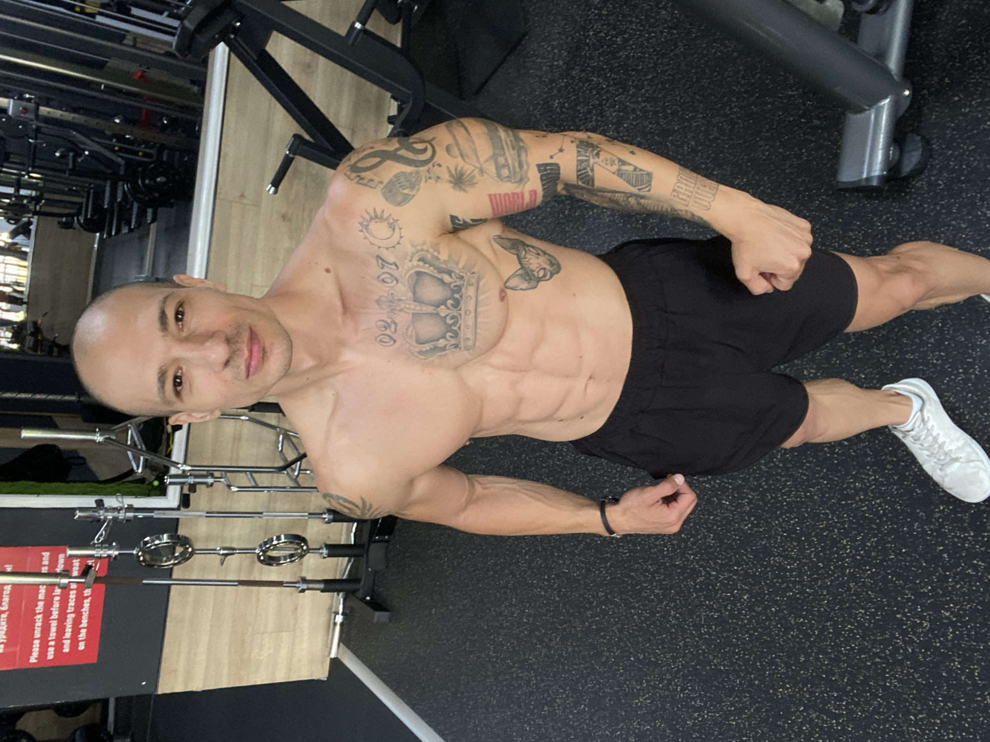The image depicts a shirtless man with visible abdominal muscles and tattoos on his chest and arm, standing in a gym. He is wearing black shorts and white athletic shoes. Behind him, gym equipment such as weight racks and benches are visible, along with a mirror reflecting more of the gym interior. A sign with text in a foreign language is mounted on the wall to the left of the man. The flooring is dark, textured, and speckled. The scene appears to be in a well-lit indoor fitness facility. This location is identified as a gym in Pleven, Bulgaria.