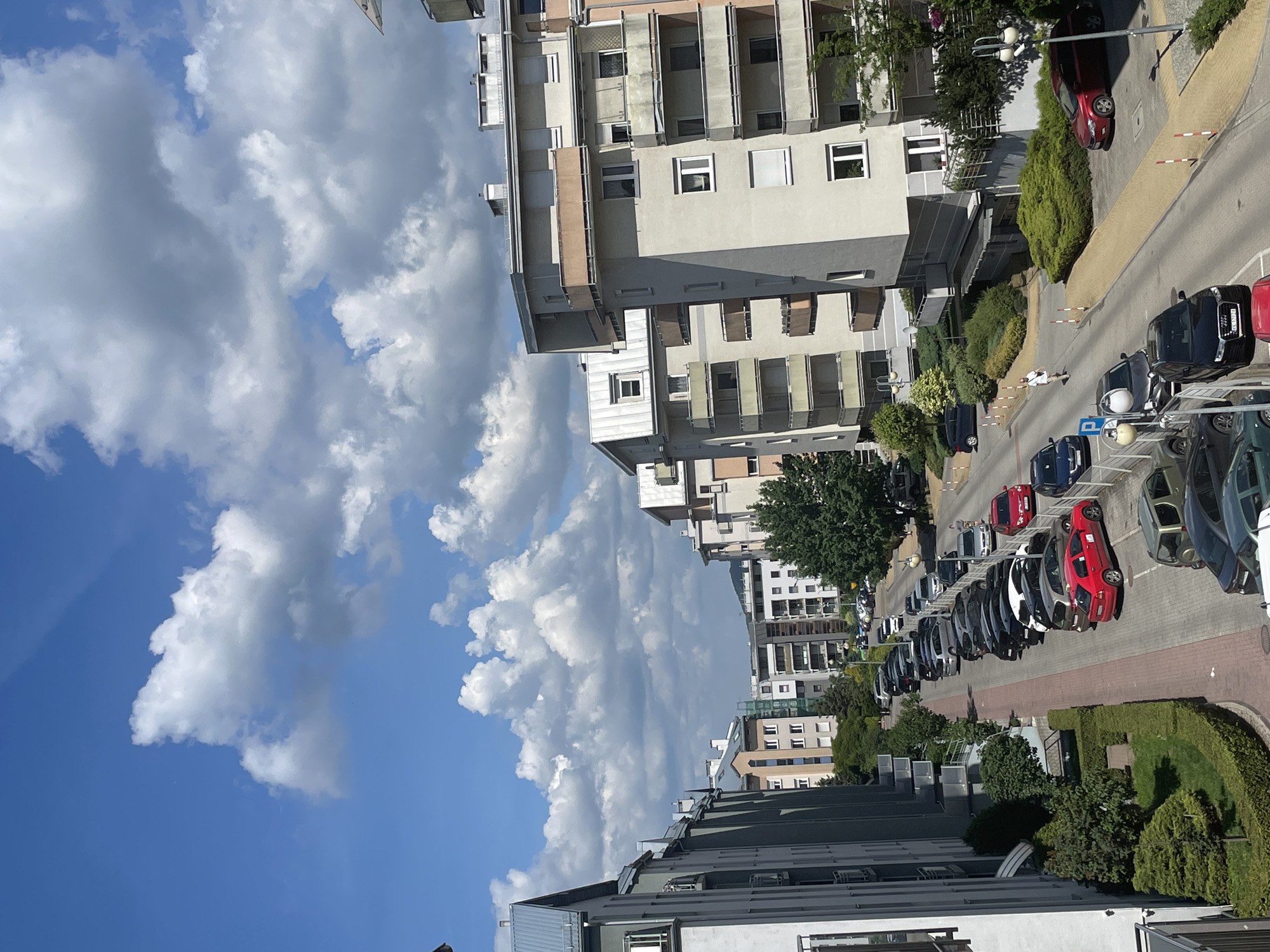 In a city setting under a blue sky with scattered white clouds, a street runs between apartment buildings and is lined with parked cars. The apartment buildings are multi-story structures with balconies and visible windows. The street is paved, with some areas marked for parking. A pedestrian is walking on the sidewalk. Various vehicles, including cars and SUVs, are parked along both sides of the street. Greenery, including trees and bushes, is visible in front of the buildings and along the side of the street. The scene appears to be a residential urban area on a clear day.