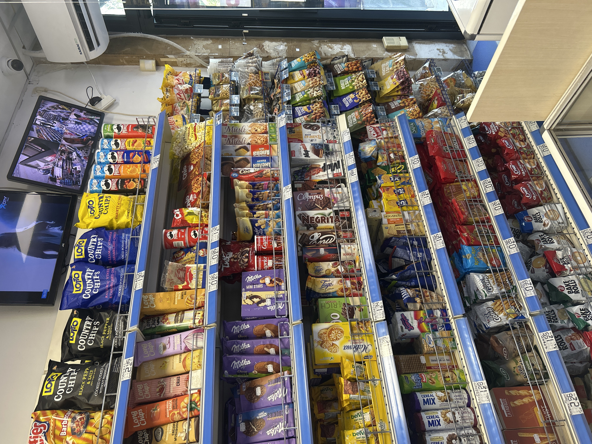 The image shows an aisle of a convenience store filled with snacks. The shelves are stocked with various chips, cookies, crackers, and candy bars. The products are mostly in brightly colored packaging. In the background, a security monitor displays multiple camera feeds of the store interior, and an air conditioning unit is mounted on the wall. The store appears to be well-lit and organized.