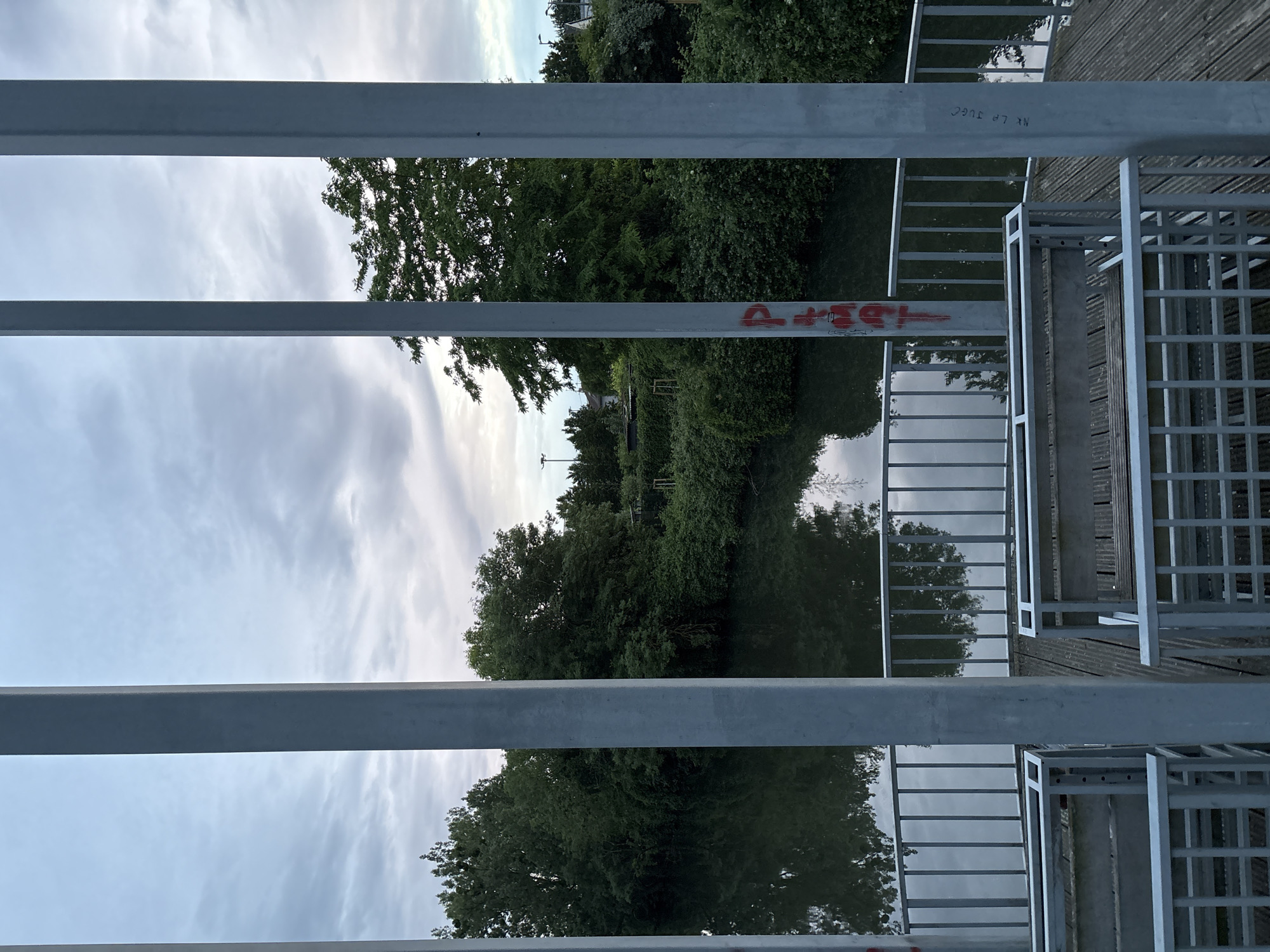 The image captures a view through metal bars, possibly a fence or railing, overlooking a body of water, likely a canal or river. The water reflects the surrounding landscape, which includes trees, vegetation, and a cloudy sky. Graffiti is visible on one of the metal bars. The location is described as the city of Unknown, Unknown. A metal structure with a grid pattern is visible on the right side of the frame, possibly a part of a platform or walkway.