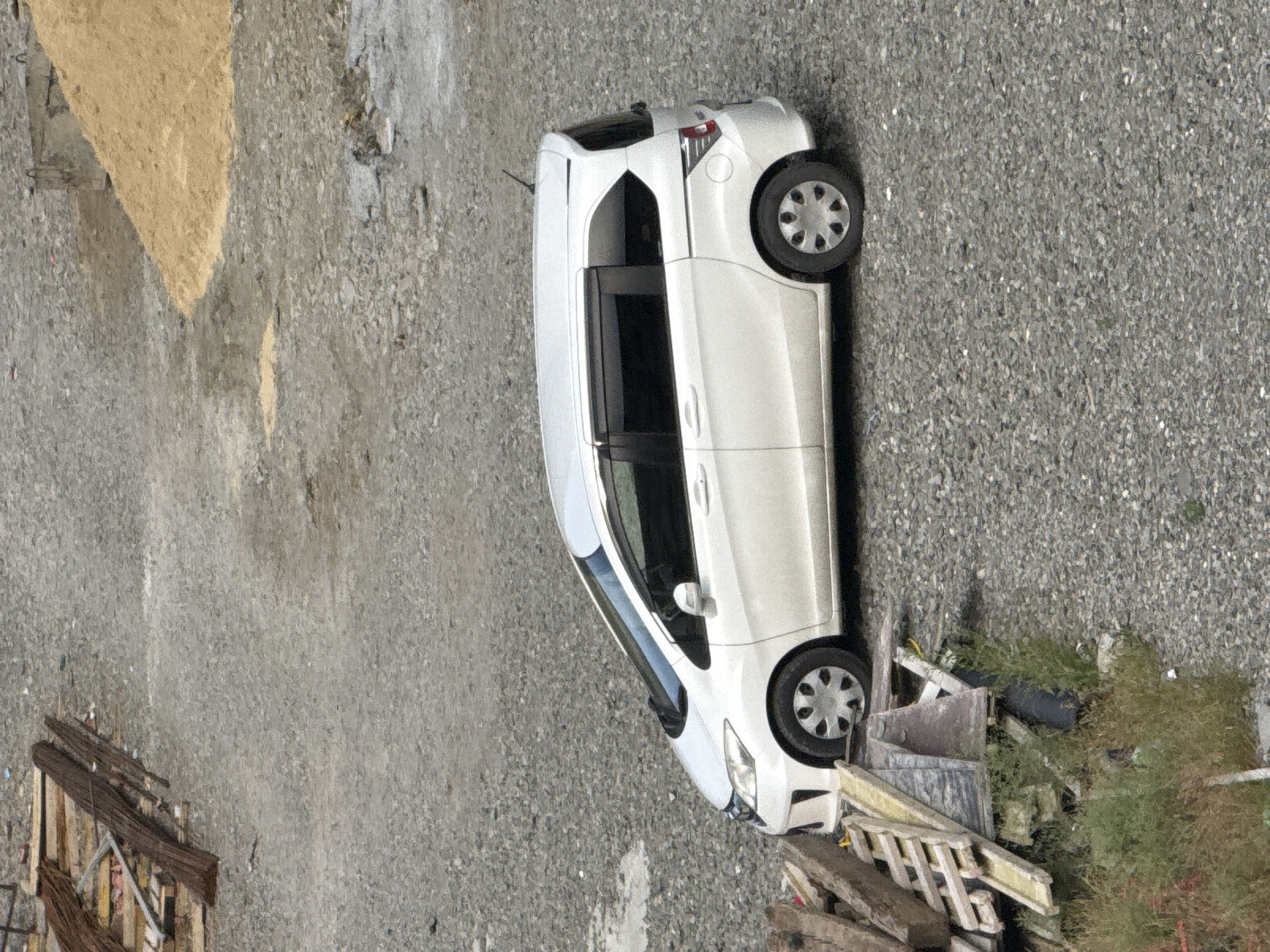 This is a top-down, rotated aerial shot of a white minivan parked on a gravel surface. The minivan is positioned diagonally across the frame. To the left of the minivan, there is an area of lighter-colored, possibly sandy or dusty ground with a rough texture. The rest of the ground is covered in small gray gravel, with some scattered debris including green foliage, wooden planks, and what appears to be construction materials. The image appears to be taken in daylight, and the weather seems clear. There is no visible text in the image. The scene suggests an outdoor storage area, a construction site, or a less maintained parking lot, possibly in Tbilisi, Georgia, given the context.