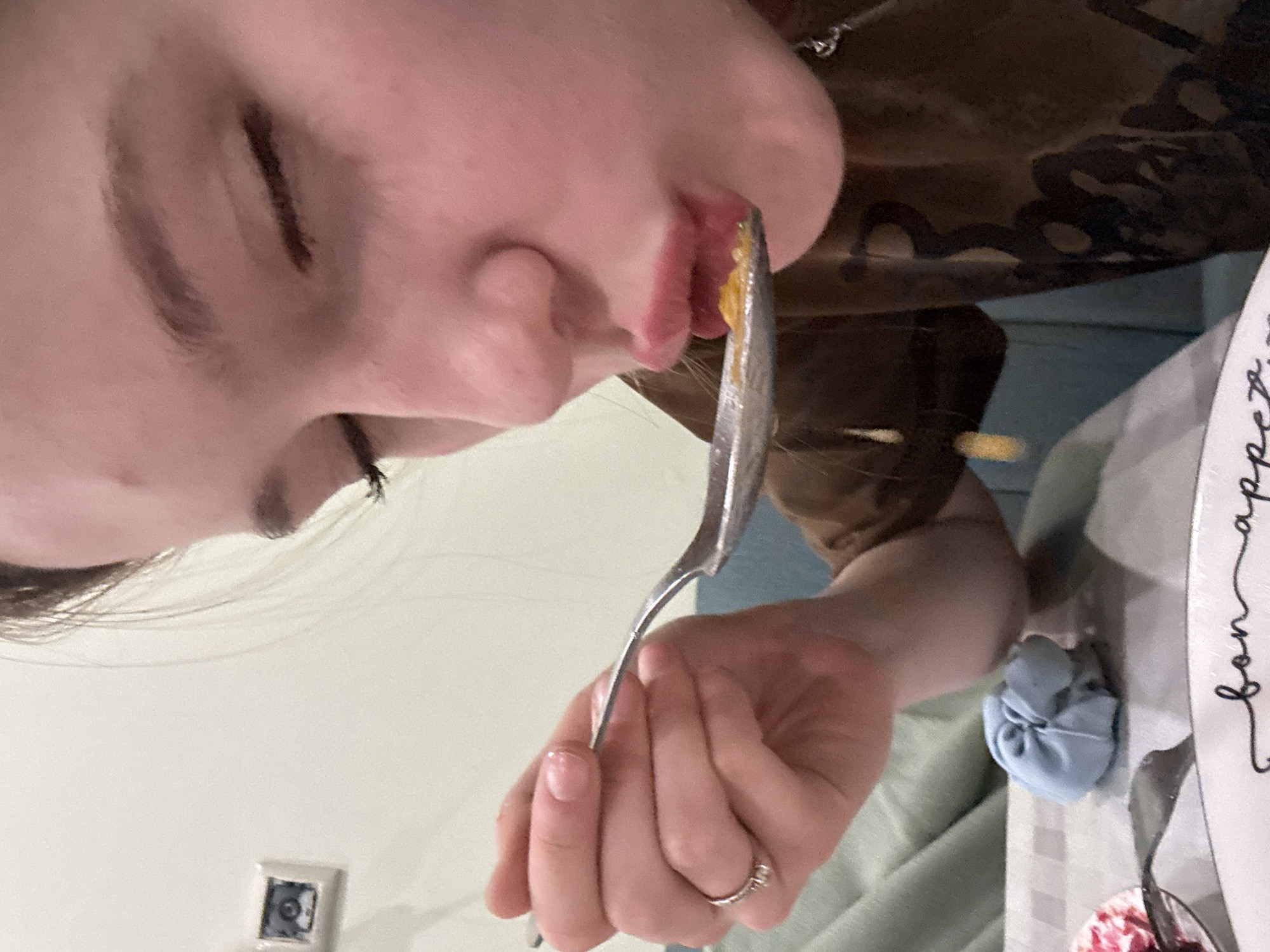 The image shows a close-up, side-profile of a young person eating from a spoon. The person's eyes are closed, and their lips are pursed as they bring the spoon to their mouth. The spoon contains a yellow, lumpy food substance, and some of it appears to be dripping. The person is wearing a brown t-shirt with a dark graphic design. Their left hand is holding the spoon, and they are wearing a silver ring on their index finger. In the background, on the wall, there is a small framed picture of an eye. On the table, there is a blue cloth object and the edge of a white plate with the words "bon appetit" written on it. The environment suggests an indoor setting, possibly a home or a casual dining establishment. There is no information available regarding the location in the city of Sinyavino, Russia.