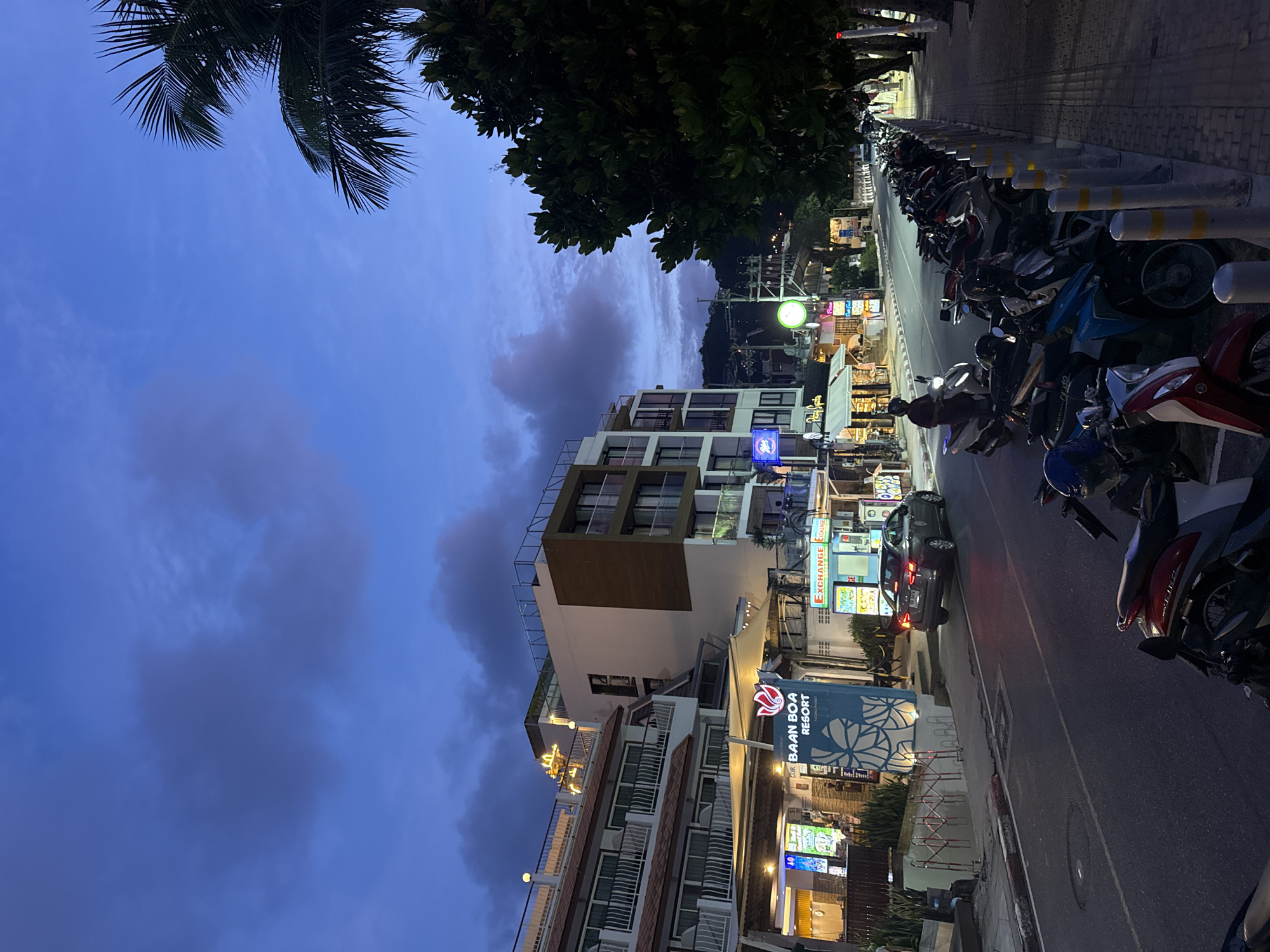 The image depicts a street scene in Patong, Thailand, at dusk or early evening. The sky is a deep blue with some scattered clouds, suggesting the transition from day to night. On the left side of the frame, the top of a palm tree and lush green foliage are visible, indicating a tropical environment.
The main subjects are buildings and a street lined with parked motorcycles. The buildings appear to be hotels or resorts, with modern architecture featuring balconies and large windows. One building prominently displays a sign for "BAAN BOA RESORT". The street itself is paved, with a pedestrian walkway alongside. The walkway is lined with bollards, some with yellow reflective bands. A significant number of motorcycles are parked along the street, indicating a popular area or a place where visitors park their vehicles.
In the background, more buildings and what appear to be shops or businesses with illuminated signs are visible. One sign clearly reads "EXCHANGE". A gray car is driving on the street, its taillights illuminated. The overall atmosphere suggests a lively tourist area, possibly a main street or a road leading to hotels and commercial establishments. The lighting suggests that it is not yet fully dark, with some artificial lights from the buildings starting to shine. No people are clearly visible in the image, but the presence of parked vehicles implies that activity is ongoing or has recently occurred. The scene evokes a sense of a warm, tropical evening in a bustling tourist destination.