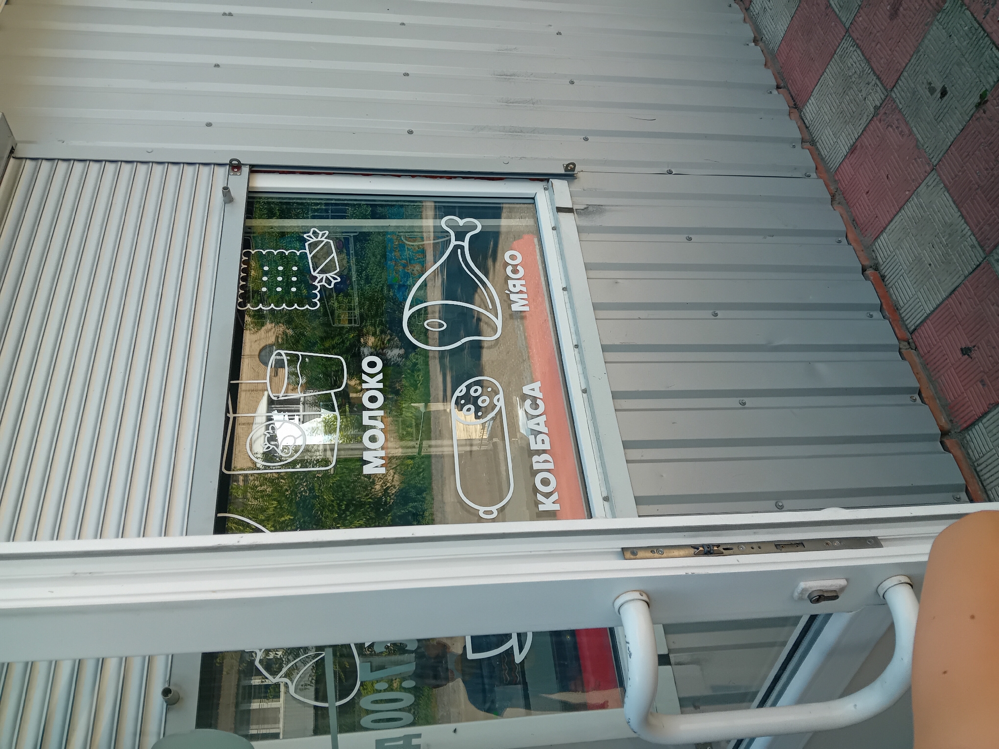 The image shows the exterior of a small retail kiosk in Chuhuiv, Ukraine. The kiosk has a gray corrugated metal exterior and a white framed glass window. White decals on the window display product icons and text. From left to right, the decals include: a clock face showing "00:15", a cartoon of a cow's head and a glass of milk with the text "МОЛОКО" (Milk), a cracker and a candy with the text "ПЕЧЕНЬЕ" (Cookies), a sausage with the text "КОВБАСА" (Sausage), and a chicken leg with the text "МЯСО" (Meat). A white handle and a lock are visible on the glass door to the left. The ground in front of the kiosk is paved with red and gray square tiles. In the reflection on the glass door, a person is partially visible, facing away from the camera.