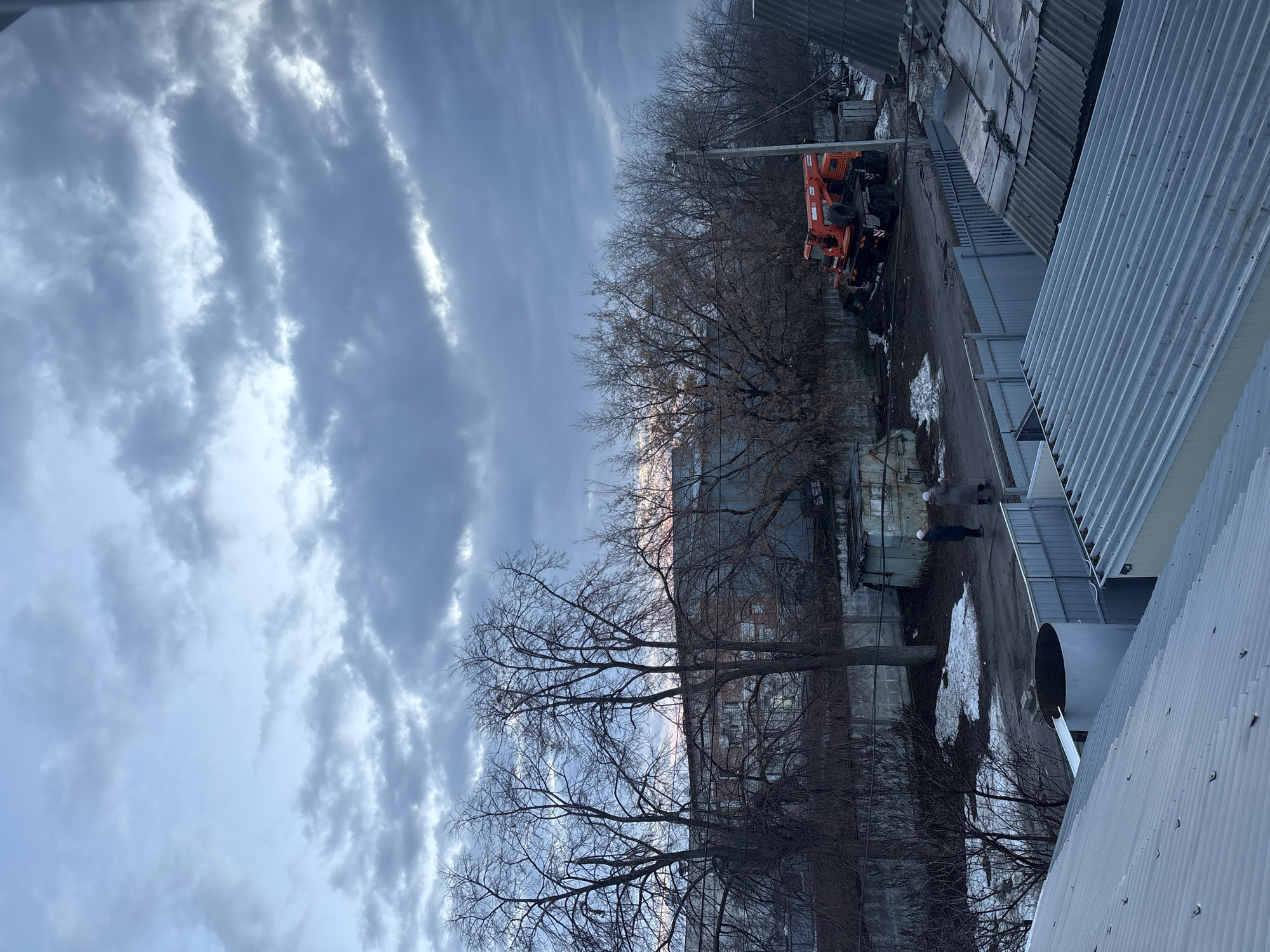 The image presents an outdoor scene in Ulyanovsk, Russia, characterized by a wintry urban environment. The sky is overcast with striated clouds. Bare deciduous trees are interspersed with industrial buildings. An orange crane is parked beside a fenced-off area. Patches of snow remain on the ground. One individual is visible, wearing a dark coat, walking along a pavement between buildings. Corrugated metal siding and roofing are prevalent on the surrounding structures.