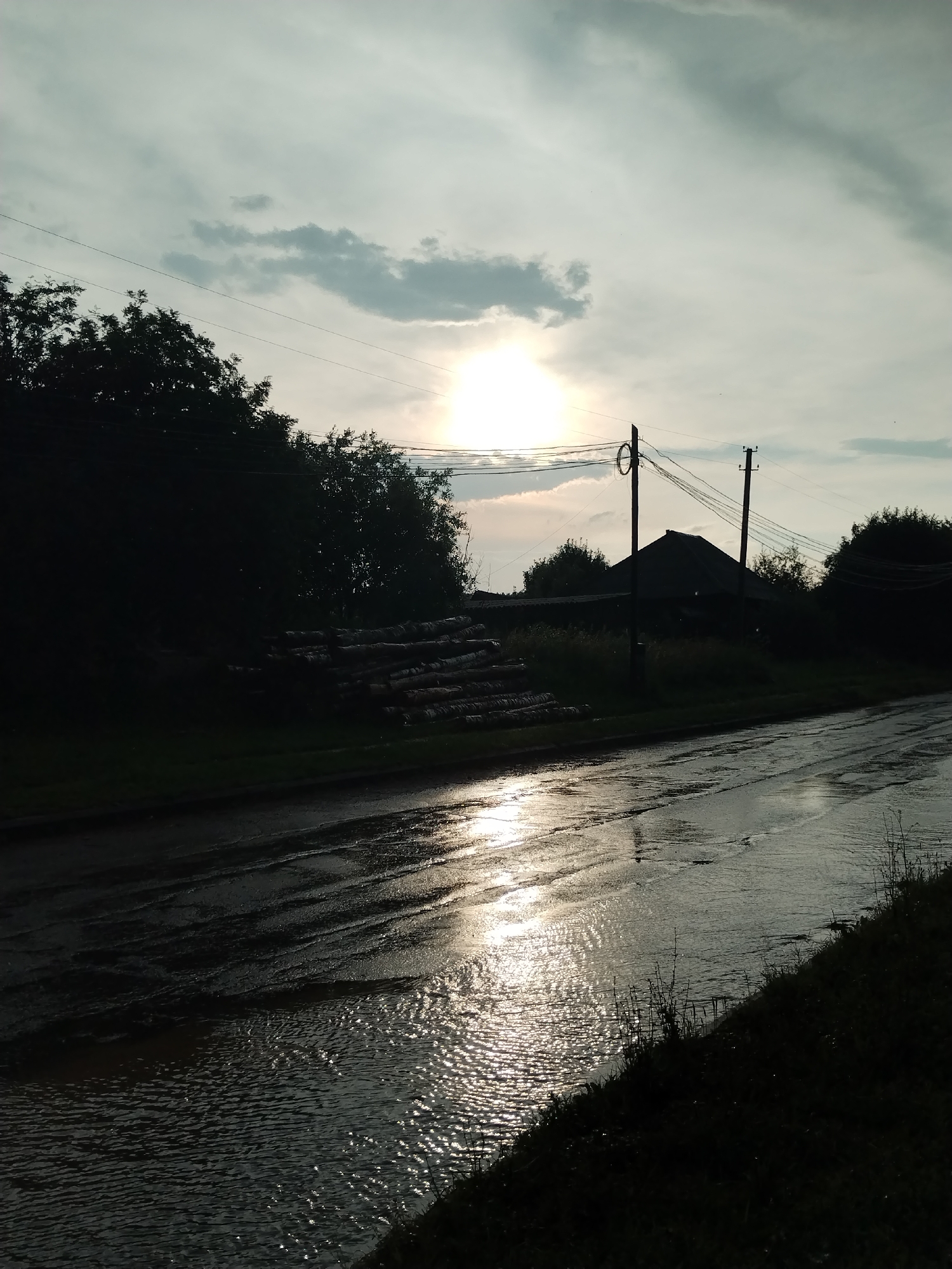 The image depicts a rural roadside scene in Sosnovo, Russia, during what appears to be overcast conditions with the sun breaking through. The foreground features a wet, muddy road with reflections of the sky and sun. To the left of the road, a pile of logs is stacked. Beyond the road, on the right side, there is a dark silhouette of a house with a steep roof, flanked by trees and foliage. Two utility poles with overhead wires are also visible in the midground. The sky is filled with clouds, with the sun positioned high and partially obscured. The overall impression is of a damp, rural landscape at dusk or dawn.