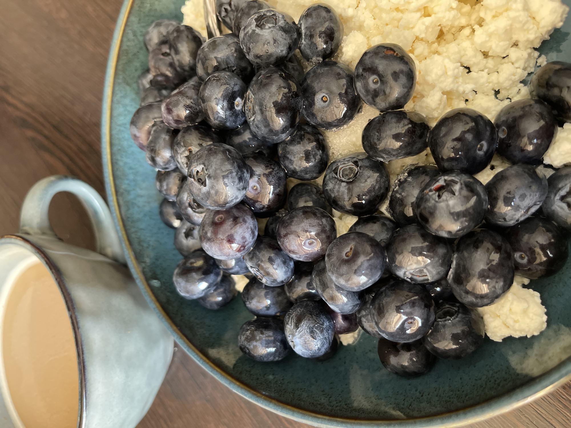 The image shows a close-up, overhead view of a rustic breakfast setting. In the foreground, a teal-colored ceramic bowl is filled with cottage cheese topped with a generous portion of fresh blueberries. The blueberries are dark purple with a matte sheen, and some appear to have a slight dusty bloom. The texture of the cottage cheese is visible, appearing lumpy and creamy. To the left and slightly behind the bowl, a matching teal ceramic mug contains a light brown beverage, likely coffee or tea with milk. The mug is partially filled, and the liquid reflects the light. The items are placed on a dark wooden surface, possibly a table. The background is blurred, suggesting a shallow depth of field, and consists of the wooden surface and the muted tones of the mug and bowl. The scene is well-lit, with natural light casting soft shadows. There are no people visible in the image. The context of Kommunarka, Russia, is not evident from the visual information in the image itself.