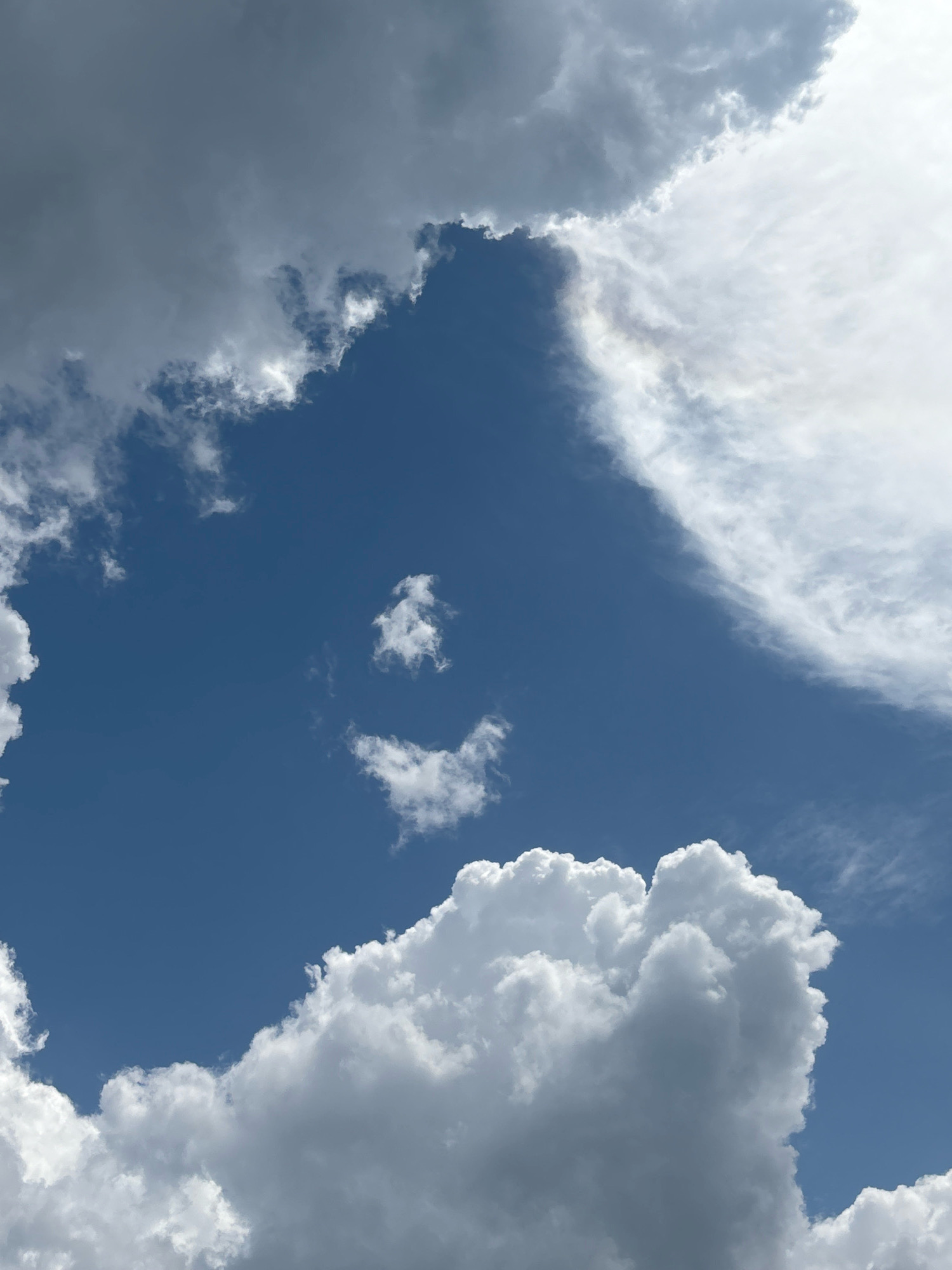 A close-up, low-angle shot captures a vast expanse of the bright blue sky above Nainpur, India, dotted with a variety of clouds. The clouds range in texture and color, from dark, brooding storm clouds on the left to wispy, white cirrus clouds on the right. In the center of the frame, two small, puffy clouds serendipitously form the shape of a smiling face, with one cloud serving as the eyes and the other as a curved smile. The sun, though not directly visible, illuminates the clouds from above, creating highlights and shadows that give them depth and dimension. The bottom of the frame is dominated by a large, cumulus cloud formation, its white peaks casting a soft glow against the blue backdrop.