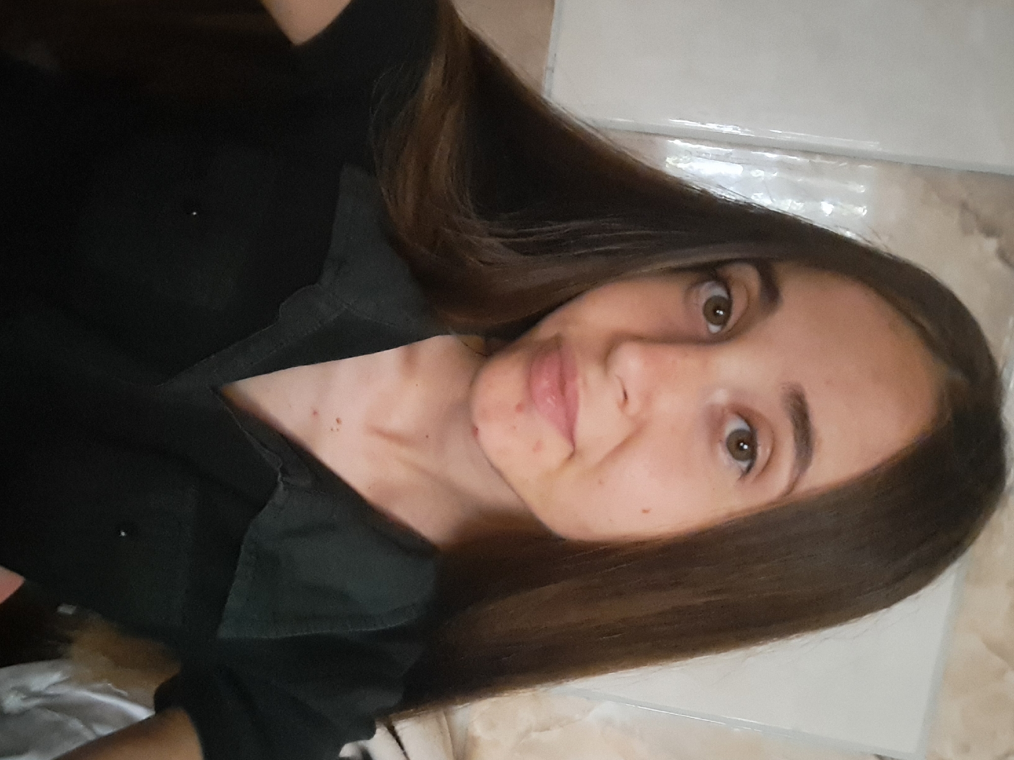 A young woman with long, dark brown hair is taking a selfie. She is wearing a dark collared shirt. The background appears to be indoors, possibly a home, with tiled or paneled walls. There are some blemishes on her face. The overall lighting suggests an indoor setting. The location is Vypasne, Ukraine.