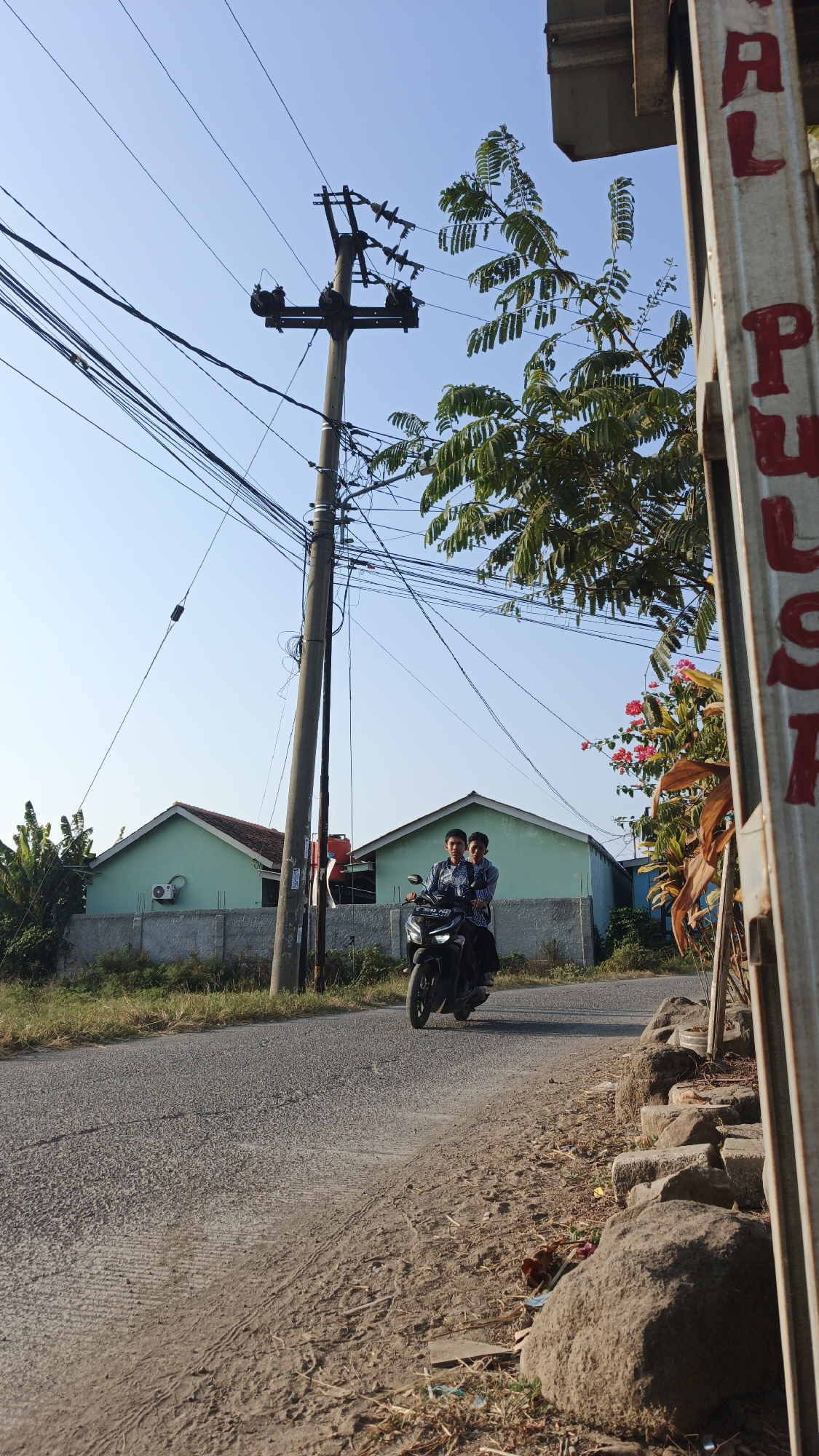 In the city of Santing, Indonesia, a scene unfolds on a paved road. Two young men are riding a black motorcycle. The rider, wearing a blue patterned shirt, is steering, while the passenger, also in a blue patterned shirt, sits behind him. They are riding away from the viewer towards a cluster of pastel green houses with dark roofs. A grey concrete wall with sparse vegetation runs along the front of the houses. A tall, weathered utility pole with numerous electrical wires is prominently positioned on the right side of the road, partially obscuring the houses. The sky above is a clear, pale blue. In the foreground, the road transitions into a dirt shoulder with scattered rocks and dry grass. To the far right of the image, a metal post with red lettering is partially visible.