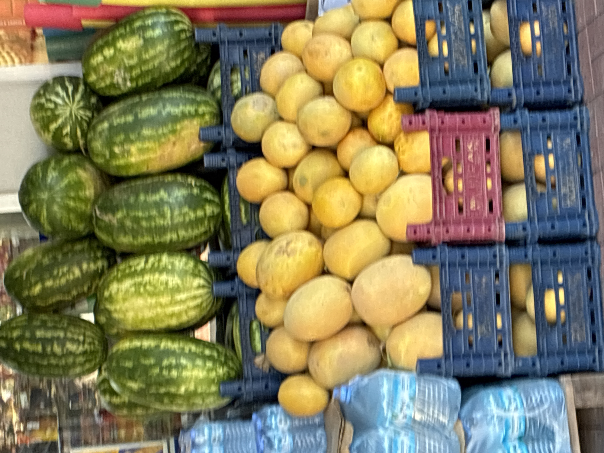 The image depicts an outdoor fruit stand in Alanya, Turkey. Prominently featured are stacks of watermelons, characterized by their dark green rind with lighter green stripes. Below the watermelons, in plastic crates, are piles of yellow-orange melons, which appear to be cantaloupes or similar varieties. To the left of the fruit are stacks of clear plastic bottles of water. The background shows glimpses of a building and other market stalls, suggesting a bustling commercial area. The overall impression is a vibrant display of fresh produce, typical of a local market.