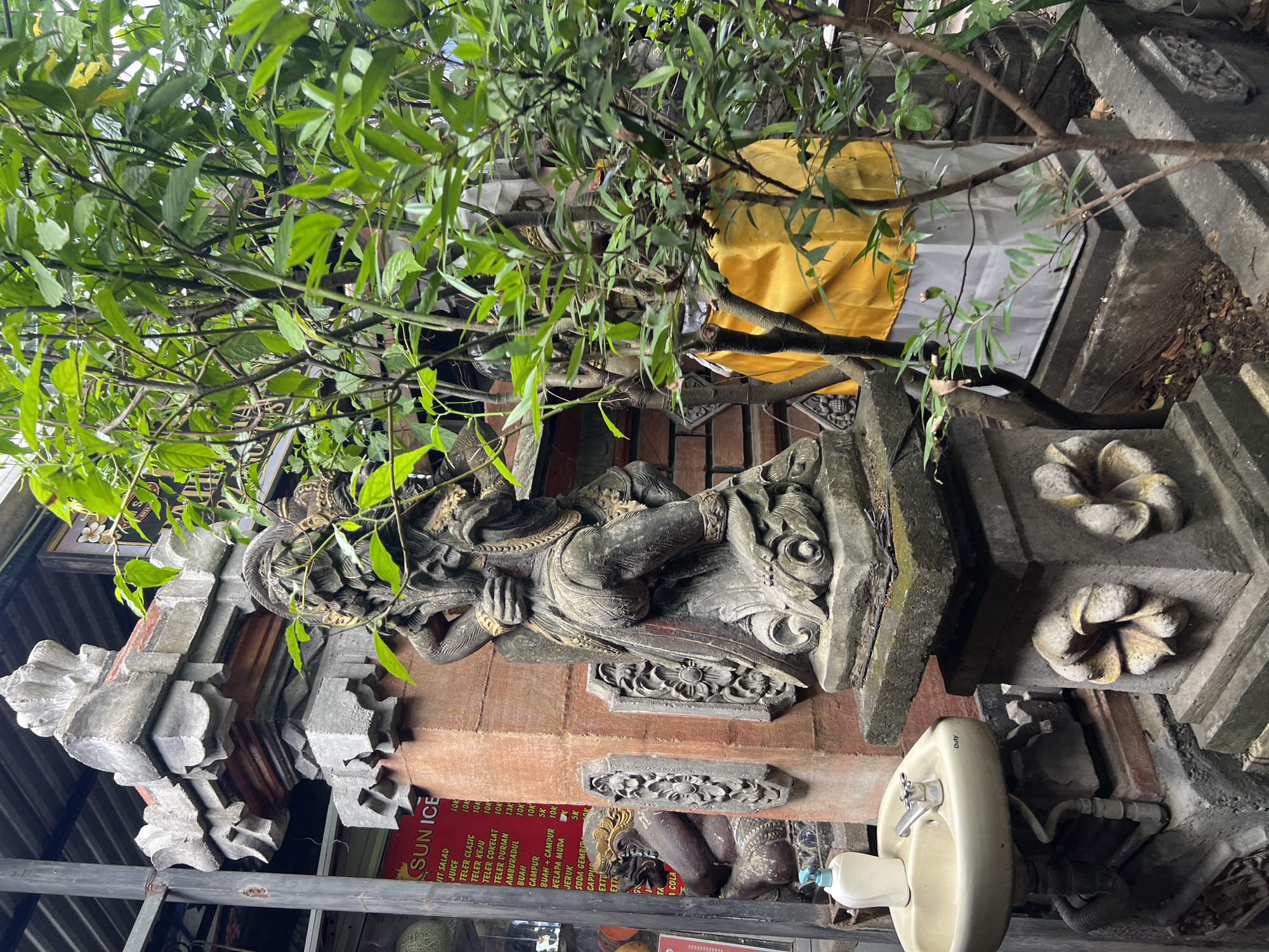 The image depicts an outdoor scene in Kuta, Indonesia, featuring traditional Balinese stone carvings and lush greenery. In the foreground, a stone sculpture of a deity or mythical figure stands on a pedestal adorned with floral motifs. To the left, a wall-mounted sink with a faucet and a pump bottle of hand soap is visible, suggesting a functional area within the outdoor space. Behind the sink, a red menu board with a list of prices in Indonesian Rupiah (indicated by "K") is partially visible, advertising items like "JUICE" and "TEA." Further back, another stone carving of a seated figure with a headdress can be seen. The background is filled with dense foliage from trees and plants, some of which are draped with yellow and white fabric, a common practice in Balinese culture to honor sacred objects or places. The overall impression is one of a cultural site or a decorative element in a Balinese garden or temple complex.