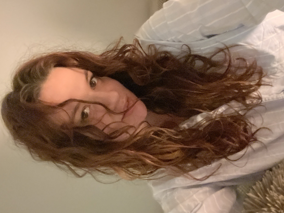 A woman with wavy brown hair is seen in a close-up shot, tilted to the side. Her face, with prominent cheekbones and a soft expression, is visible amidst the curls. She has light skin and brown eyes that are looking toward the camera. She's wearing a light blue and white striped shirt, which is casually draped around her. The background appears to be a plain, light-colored wall.