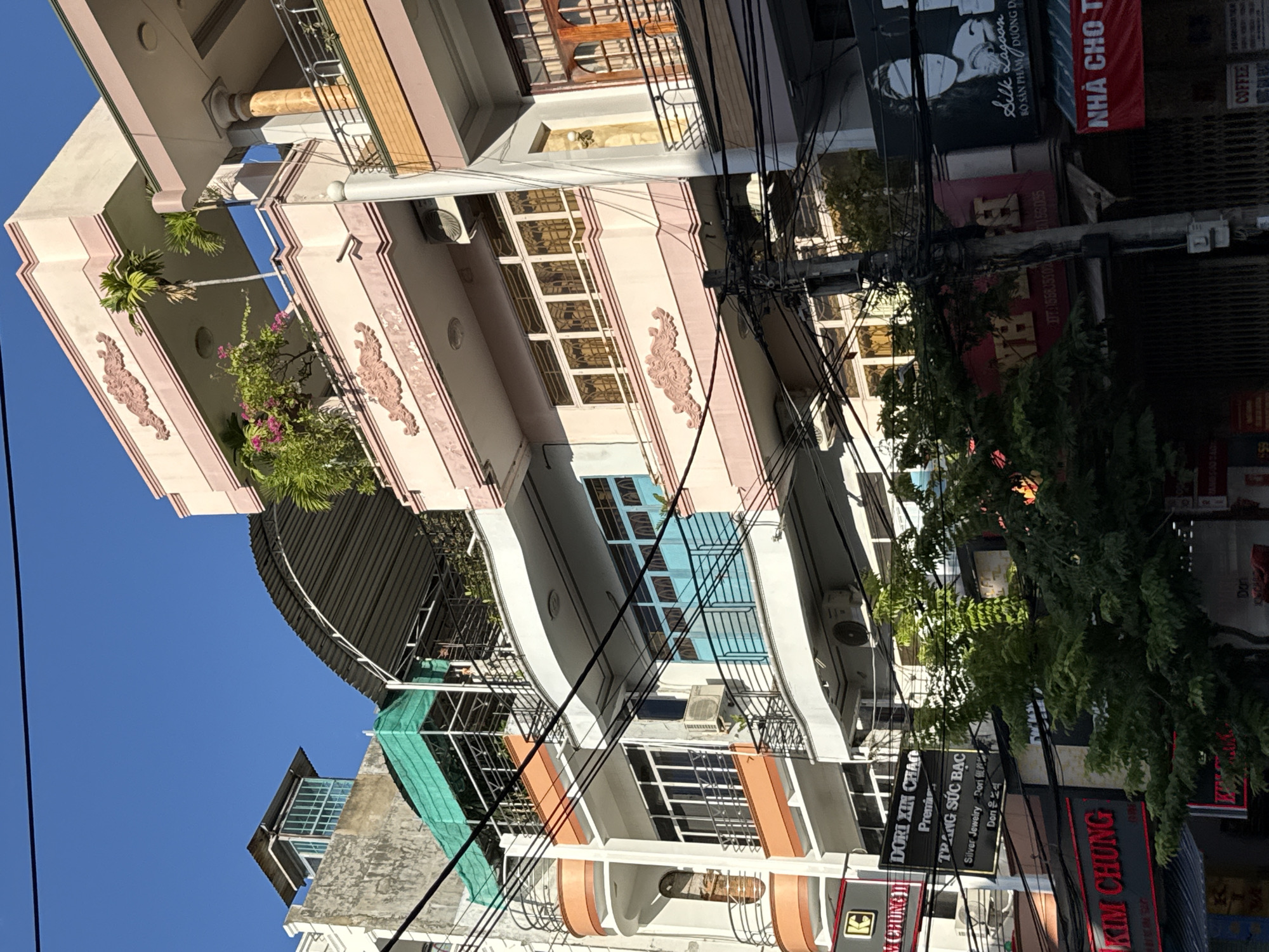 This is a street-level, eye-level, outdoor, daytime, medium shot of a row of multi-story buildings in Nha Trang, Vietnam. The buildings are a mix of architectural styles, with some featuring modern balconies and others with more traditional detailing. Many of the buildings are painted in pastel colors like pink and orange. Electrical wires are strung across the foreground, obscuring some of the buildings and creating a sense of urban density. There are several signs visible, including one that reads "DORI XIN CHAO" and "TRANG SUC BAC," suggesting shops selling silver jewelry. Another sign displays the words "KIM CHUNG" in red illuminated letters, likely a business name. Trees with lush green foliage are growing in front of some of the buildings, adding a touch of nature to the urban landscape. The sky is clear and blue. No people are prominently visible in the frame.