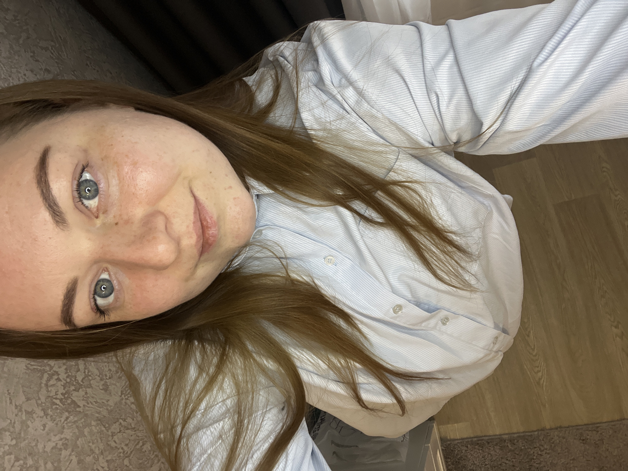 A young woman with light brown hair and fair skin is centered in the image, smiling slightly. Her light blue eyes are visible. She is wearing a light blue striped button-down shirt. The background includes a section of light brown wooden flooring, a grey upholstered surface, and dark curtains or fabric in the top left corner. There is no information to indicate the location is in the city of Раздолье, Russia.