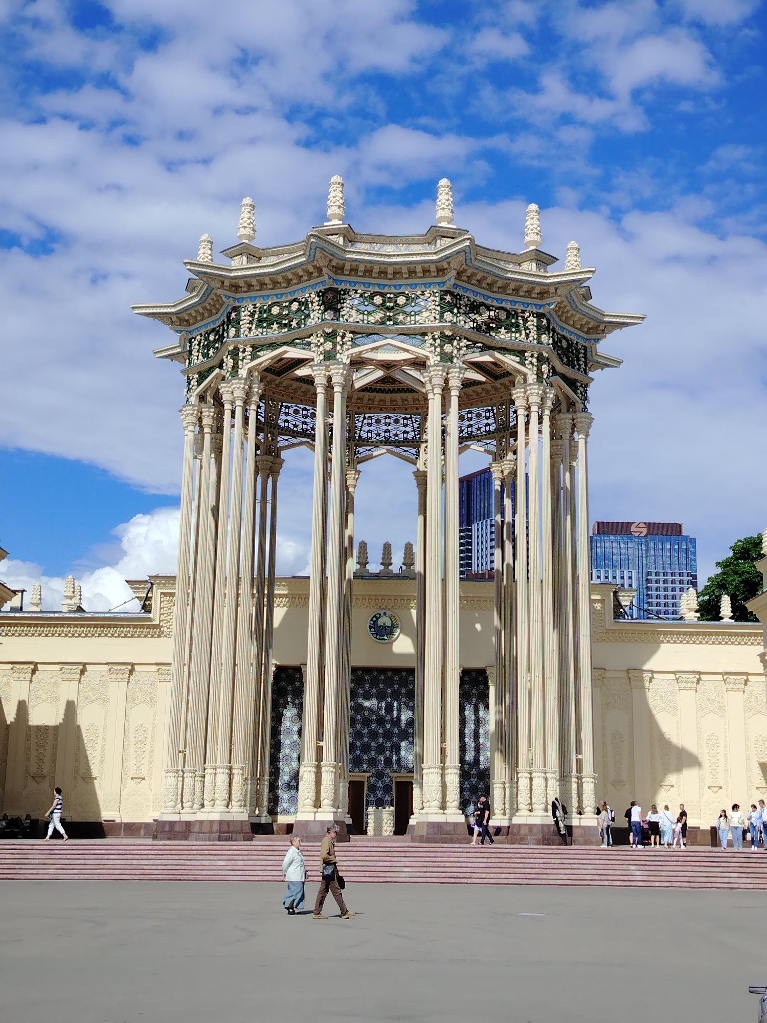 This is a full-frame, eye-level, outdoor, vertical shot of the "Friendship of Nations" fountain pavilion at VDNKh (Exhibition of Achievements of National Economy) in Moscow, Russia. The pavilion, a prominent Soviet-era monument, is elaborately decorated with gilded elements and intricate patterns, featuring numerous tall, fluted columns supporting a tiered, multi-story roof structure topped with decorative spires. The pavilion is cream-colored with blue and green accents on the roof.
In the foreground, a paved plaza is visible, with a few people walking across it. Two individuals, a man and a woman, are walking towards the left side of the frame. Further back, on the wide, multi-tiered steps leading up to the pavilion, a group of people are gathered and walking. One person in a striped shirt is walking alone up the steps on the left side of the frame.
Behind the pavilion, modern high-rise buildings, one with a red logo at the top and another with blue glass facades, are visible against a bright blue sky with scattered white clouds. The sun casts shadows on the plaza and the facade of the pavilion, indicating it is daytime. The overall scene depicts a sunny day at a public exposition site.