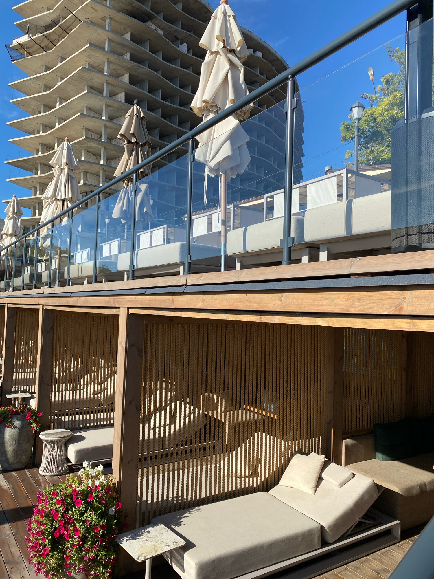 This image captures a resort area in Odesa, Ukraine, likely near a construction site for a new building. In the foreground, there are several sun loungers with beige cushions and pillows, arranged under wooden beams and a rope-like screen. One lounger is angled, and some have small side tables. Pink and red petunias are in a pot on the left, and a round, woven table sits nearby. The middle ground features a glass-paneled balcony with closed, beige umbrellas and a metal railing. Beyond this, a multi-story, unfinished concrete building dominates the background, with its skeletal structure visible against a clear blue sky. In the distance, trees and a street lamp are also visible. The lighting suggests it is daytime, with sunlight casting shadows.