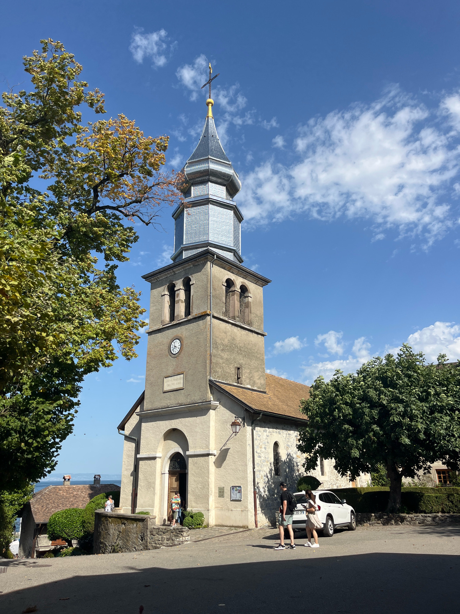 This is a medium-shot, eye-level, outdoor photograph taken on a sunny day in Yvoire, France. The photograph features a church with a tall bell tower and a copper spire topped with a cross. The church is made of stone and stucco, with arched windows and a clock on the tower. A tree with green and yellow leaves is in the foreground on the left, and another tree with green leaves is on the right. There are two people standing near a white car in front of the church. A third person, wearing a colorful shirt, is standing at the church entrance. In the background, there are other buildings with tiled roofs and a body of water, possibly a lake. The sky is blue with scattered white clouds.