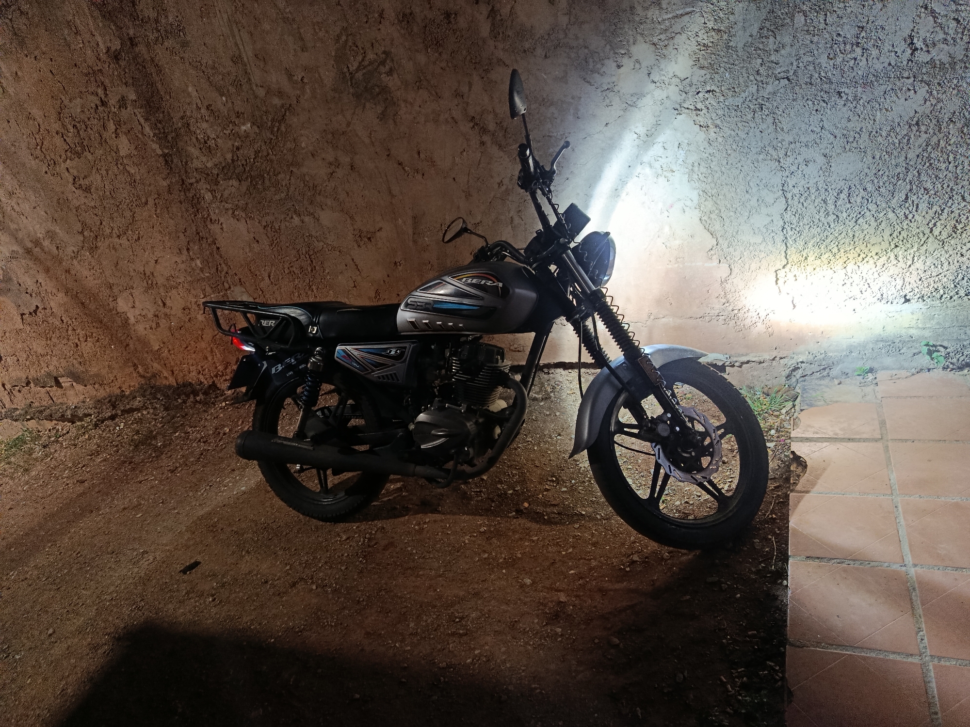 An image captured in Juan Griego, Venezuela, displays a grey Bera 150 motorcycle parked outdoors at night or in low light conditions. The motorcycle is positioned facing right, with its headlight actively illuminating a textured, rough wall to its right. The wall exhibits varied tones, transitioning from a darker reddish-brown on the left to a brighter, light grey where the headlight beam directly strikes it. The ground beneath the motorcycle consists of a reddish-brown dirt or gravel path on the left and a terracotta-colored tiled patio or walkway on the right, separated by a distinct edge. The motorcycle features a grey fuel tank with blue, silver, and black graphic decals, a black seat with a rear cargo rack, and a visible engine assembly. Its front wheel is equipped with a disc brake. Shadows from the motorcycle are cast on the dirt ground in the foreground. No individuals are present in the scene.