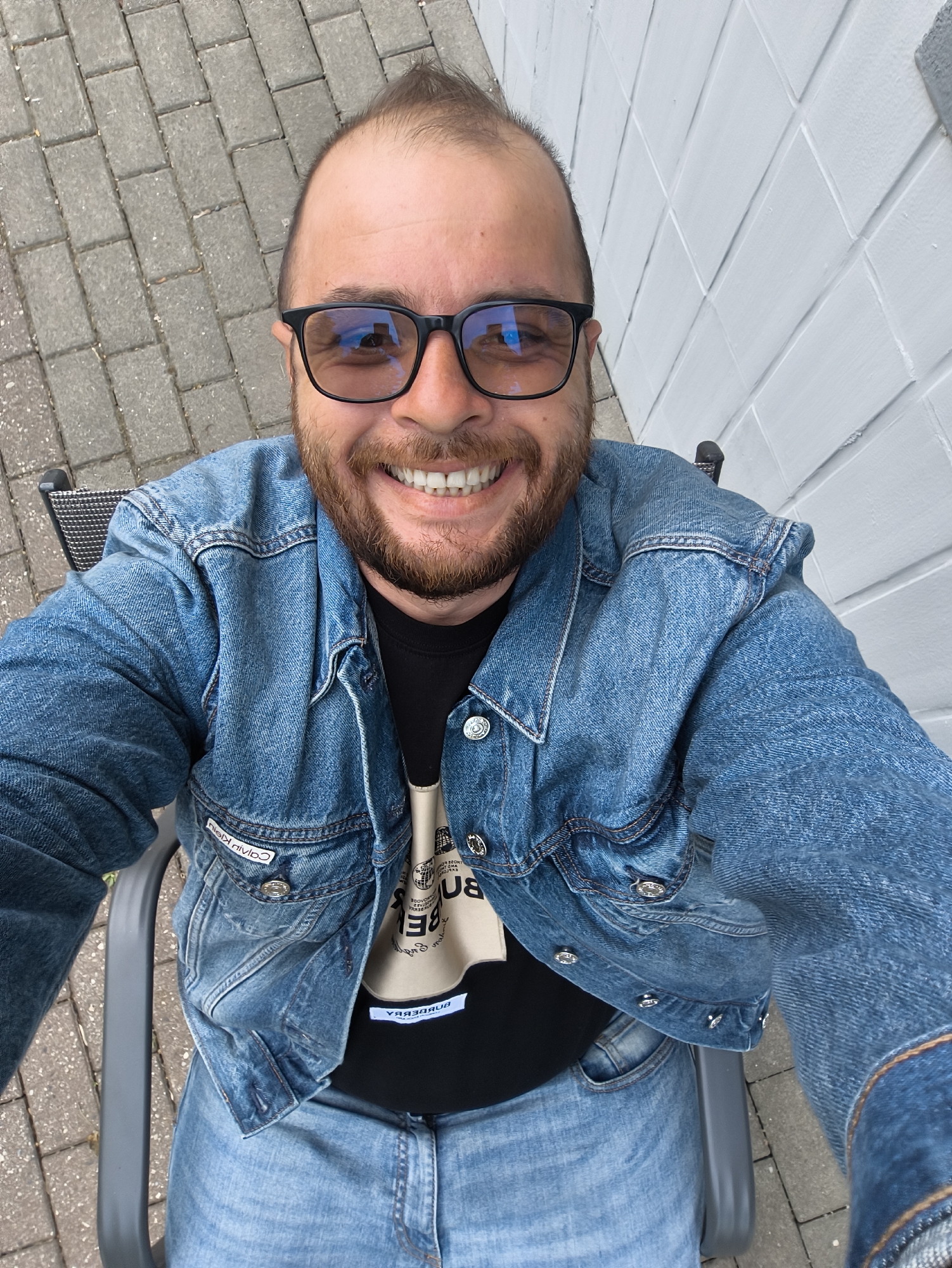 A man is sitting in a gray metal chair outdoors. He is wearing a denim jacket over a black t-shirt, denim jeans, and black framed glasses. He is smiling. The ground is paved with gray bricks. Behind him is a white painted brick wall. The location is Unknown, Unknown.