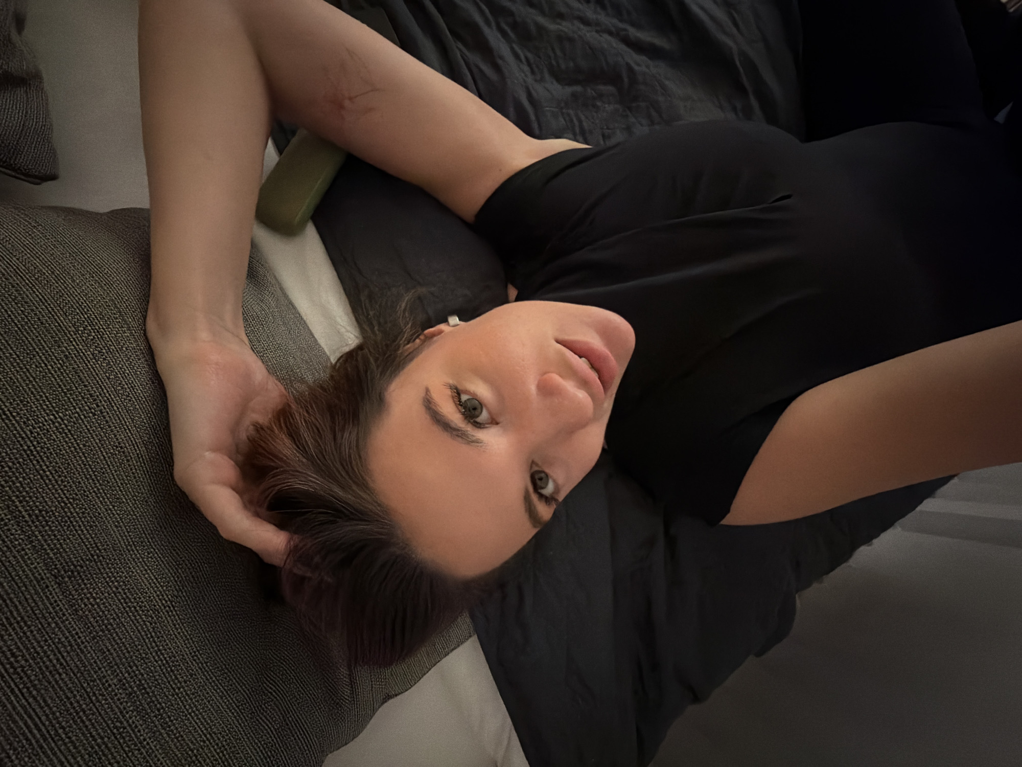 A female individual is depicted lying on her back on a bed in an indoor setting in Belgrade, Serbia. She has fair skin, dark brown hair, and green eyes, and is wearing a black short-sleeved top. Her head is angled towards the camera, with her gaze directed forward. Her lips are slightly parted. A small, silver, square-shaped earring is visible in her left ear. Her left hand is positioned behind her head, touching her hair. Red linear marks are visible on her left forearm. Her right arm rests by her side. The bedding consists of dark, likely black, sheets or a duvet, and a textured grey pillow or headboard is behind her. A white pillow or sheet is partially visible near her head on the right. A small, light green, rectangular object is positioned beside her right arm. The scene is illuminated by an immediate light source, indicating low ambient lighting conditions.