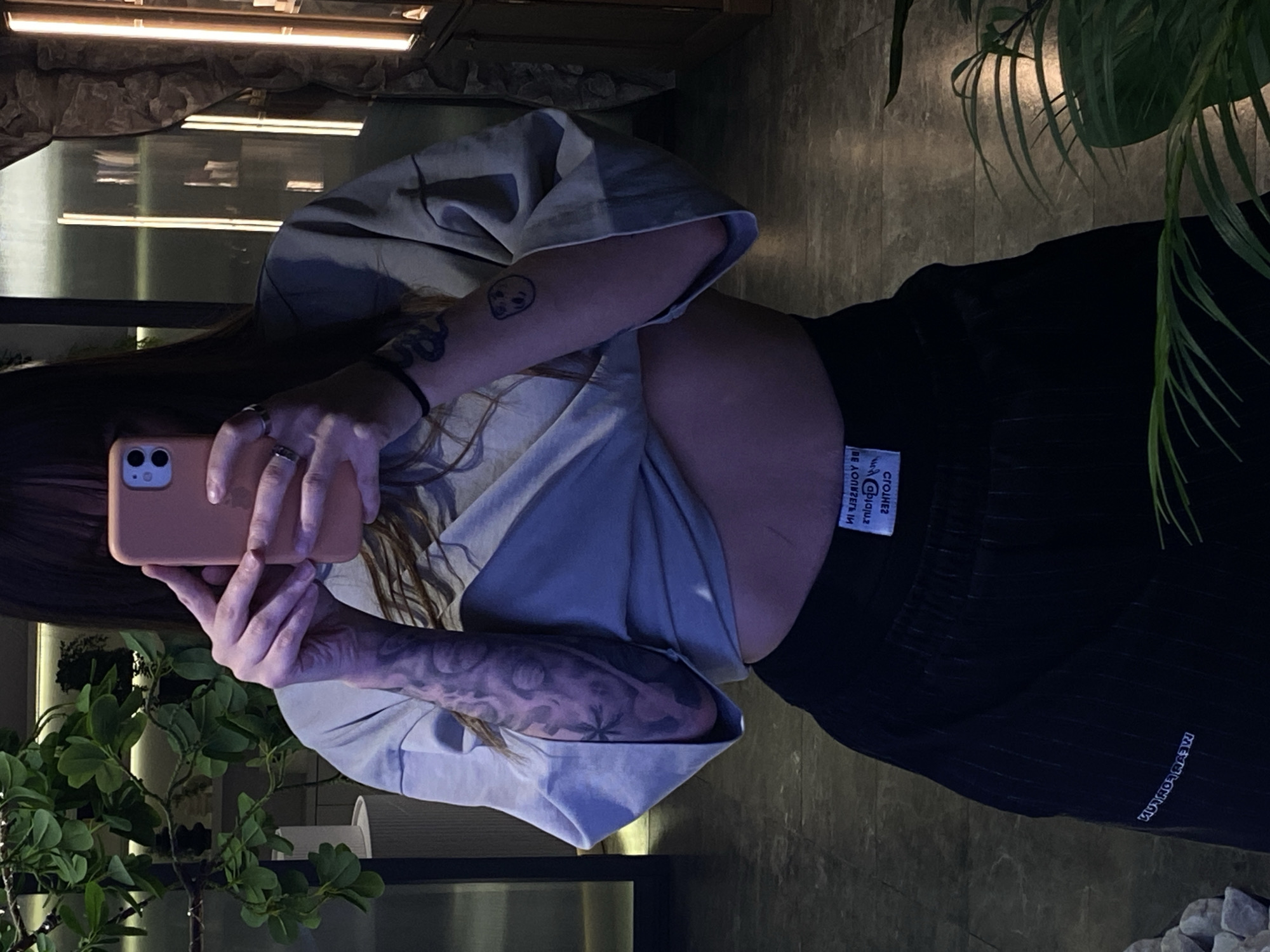 A woman is indoors, holding a peach-colored smartphone to take a photo or video. She wears a light blue, short-sleeved shirt with a dark, possibly black, skirt or pants. Tattoos are visible on both forearms, one showing various shapes and patterns, and another, smaller tattoo on the right upper arm that appears to be a simple face. A black band is on her wrist. A plant with green leaves is situated to her left. Other plants can be seen in the background, as well as what appears to be a decorative fish tank or terrarium, with a light above. The picture was taken indoors. There is no available information to indicate this image was taken in the city of Votkinsk, Russia.
