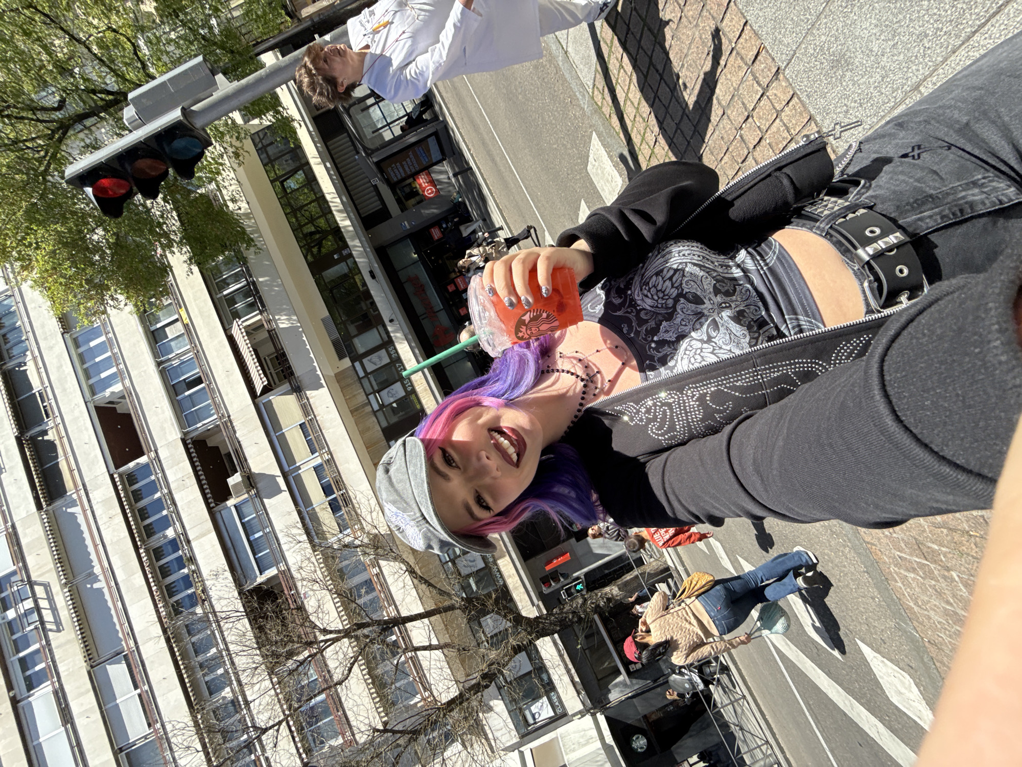 A selfie is captured outdoors in Madrid, Spain, during daylight. The subject, a young woman with pink and purple hair wearing a gray cap, black top, studded belt, and jeans, holds a Starbucks beverage. The background features a street scene with a tall building across the street and a traffic light showing a red light. Other pedestrians are present, with one wearing a white lab coat. Sidewalk brick and street markings are visible.