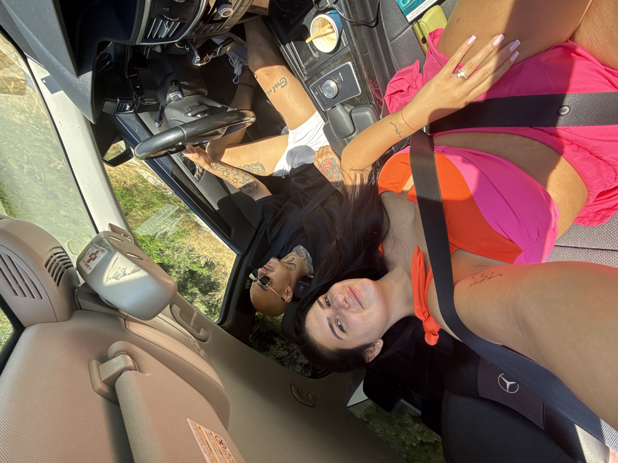 In the interior of a car, two people are visible. A woman in the foreground is wearing a brightly colored bikini and a seatbelt. She is smiling and looking at the camera. Behind her, a man with a shaved head and sunglasses is driving. He has tattoos on his arm and neck. The car appears to be parked or moving slowly on a road surrounded by vegetation. The sunlight is strong, casting shadows within the car. There are also visible car interior elements such as the dashboard, steering wheel, and air vents. The background appears to be a natural, possibly rural environment with trees and dry grass.