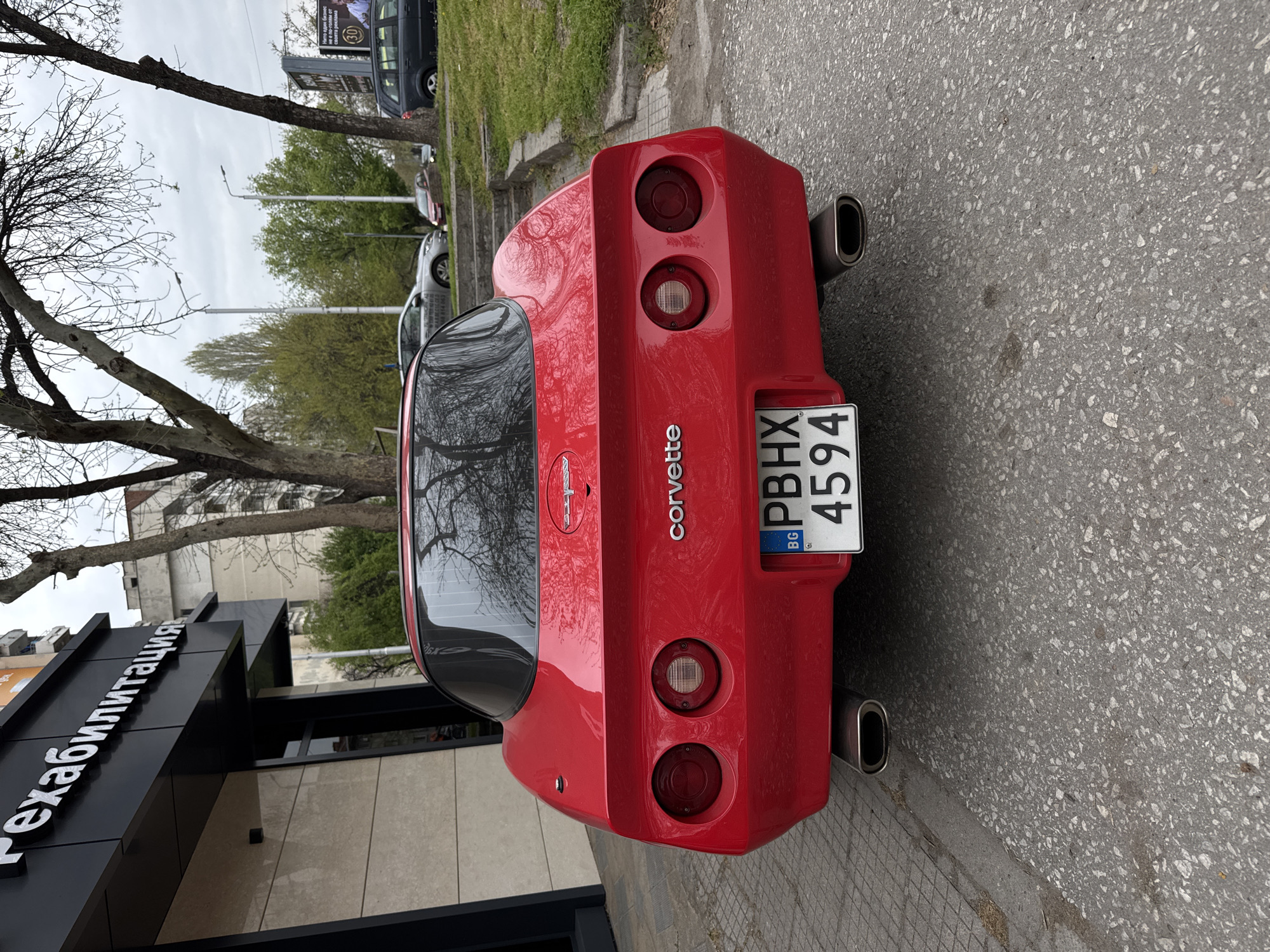 The image shows the rear of a red Corvette C4, parked on the street in Plovdiv, Bulgaria. The license plate reads "PBHX 4594." The building "Рехабилитация" is visible on the left. The background includes trees, buildings, and other parked vehicles. The sky is overcast.