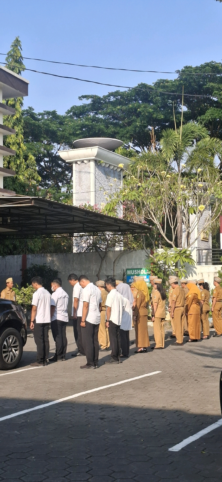In Gumulan, Indonesia, a group of people in uniform stand in a parking lot outside a building. The building appears to be a government or official institution, with a sign that reads "MUSHOLLA" visible on the wall. The people are divided into two groups: one group wears white shirts and dark pants, while the other group wears khaki-colored uniforms with matching hats. The khaki-clad individuals appear to be women, based on their attire and hairstyles. They are standing in a line, facing away from the camera. A black car is parked to the left of the frame. The environment is sunny, with trees and clear blue sky visible in the background. The ground is paved with interlocking hexagonal stones.