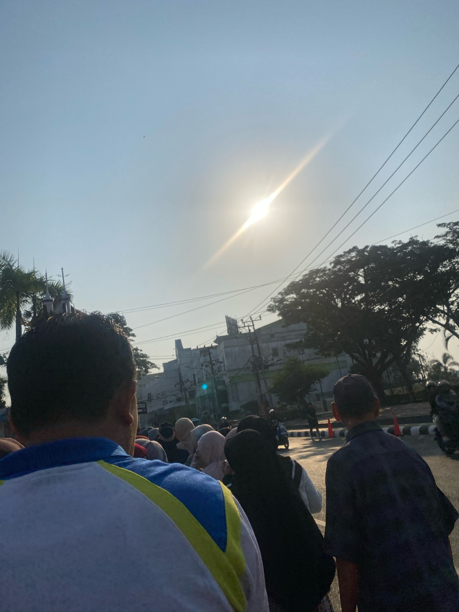 The image depicts a street scene in Banjarmasin, Indonesia, under a bright, sunny sky. The sun is positioned high in the sky, casting a strong glare and creating a sun flare that extends diagonally across the frame. Several individuals are visible, seen from behind, walking or standing on what appears to be a road or sidewalk. They are dressed in casual attire, with some wearing headscarves. Palm trees and other foliage are present, adding to the tropical environment. In the background, buildings, possibly part of a commercial or administrative area, are visible, along with power lines stretching across the sky. The road itself has traffic cones and a few motorbikes, suggesting moderate activity. The overall impression is of a normal day in a bustling city.