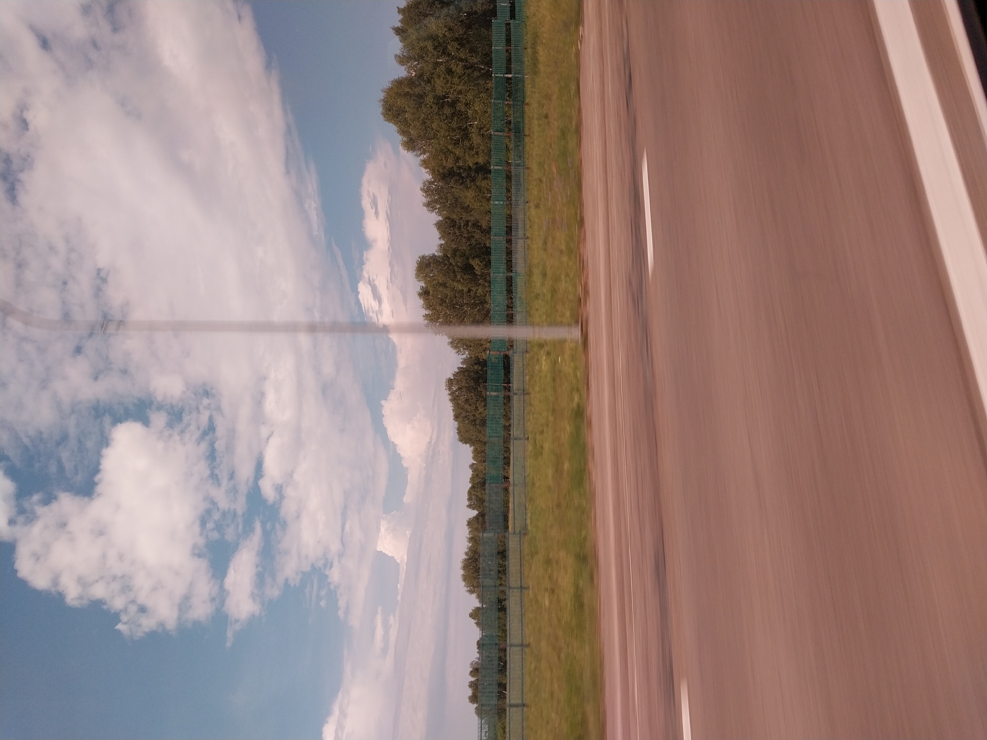 This is a photograph taken from a vehicle moving along a road in Nalobikha, Russia. The image is oriented vertically, with the right side of the road occupying the majority of the frame. The road surface is a light brown color and appears to be asphalt, with two white lane lines visible. The motion blur indicates the vehicle is in transit.
To the left of the road, a strip of green grass is visible, bordering a tall, green, chain-link fence. Beyond the fence, a dense line of trees forms a backdrop. The trees are a mix of leafy deciduous trees, appearing green and full, suggesting a summer or late spring setting.
Above the trees, a vast sky stretches out, filled with a mix of white, puffy clouds and streaks of lighter cirrus clouds against a bright blue background. The lighting suggests it is daytime, likely late morning or afternoon, with clear, fair weather. There is no visible text in the image. The scene evokes a sense of travel and the open road, with a peaceful, natural landscape to the side.