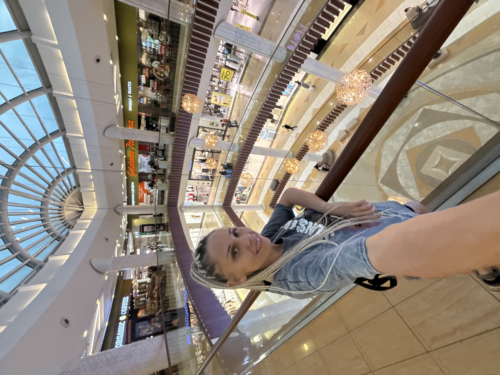 A woman with blonde braided hair, wearing a grey t-shirt, takes a selfie in a multi-level shopping mall. The mall has a large, arched glass ceiling. Escalators and walkways connect the different floors, which are lined with shops. Decorative spherical lights hang from the ceiling. In the background, shoppers can be seen browsing in stores and walking on the various levels. The woman is leaning on a railing, looking at the camera with a smile. The mall appears to be located in the city of Arkhangelsk, Russia, indicated by the text on some of the shop signs.