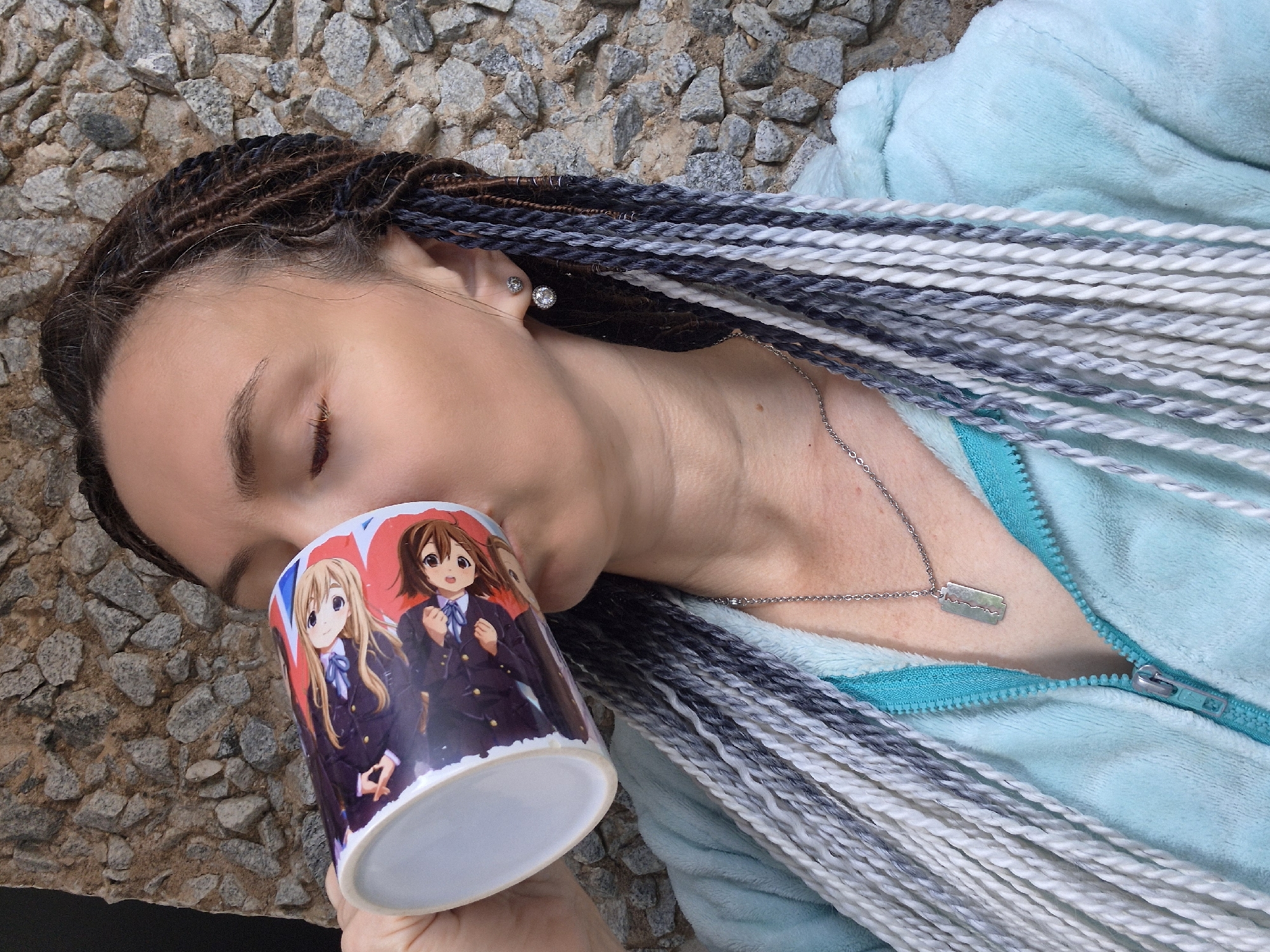 In this close-up, medium shot, a person with dark hair braided with white and grey extensions drinks from a mug featuring cartoon characters. The person is wearing a light blue hooded jacket with a turquoise zipper and a silver necklace with a rectangular pendant. The background consists of a rough, grey stone wall. The lighting suggests it is daytime. No specific location in Chaikovsky, Russia, is identifiable from the visual information.