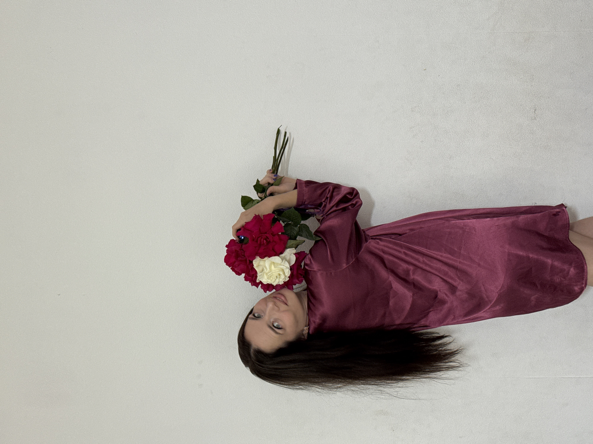 A woman with long, dark hair is posing against a white, textured wall, holding a bouquet of red and white flowers. She is wearing a burgundy satin robe. The lighting is even, casting no significant shadows. There is no discernible indication of the location being in Rusalevka, Russia, nor are there any elements within the image that point to a specific city or country.