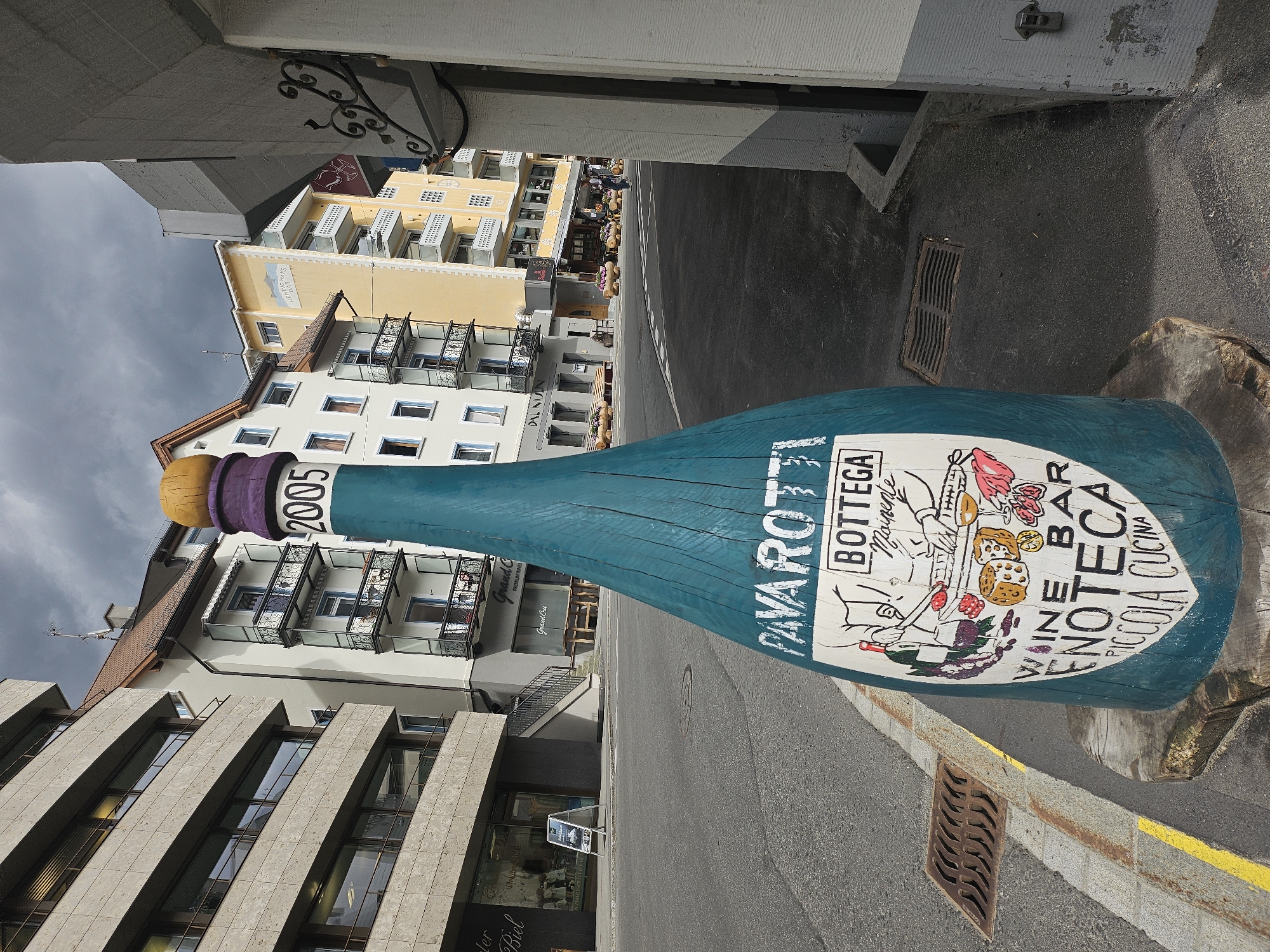 This is a photograph taken in St. Moritz, Switzerland, featuring a large, teal-colored wooden sculpture of a wine bottle, lying on its side on a street. The bottle is decorated with colorful illustrations and text. The text visible includes "2005", "PAVAROTTI", "BOTTEGA MAISONNE", "WINE BAR", "VENOTECA", and "PICCOLA CUCINA". The sculpture is situated on a street with a yellow painted line and a metal grate.
Behind the sculpture, several buildings are visible. The architecture suggests a European setting, with balconies and windows characteristic of alpine towns. One building is painted yellow and has the text "HEMMIG WAYS" and "Grand Cru" visible. Another building is lighter in color, with multiple floors and balconies. The overall scene appears to be a street in a town, possibly a commercial area given the signage on the buildings and the presence of an advertising sign in front of a store on the left.
The sky is overcast and appears to be a cloudy day. The lighting suggests it is daytime, but the weather might be cool or about to rain. There are no people prominently featured in the image, though there might be a few figures in the background of the yellow building. The focus is on the wine bottle sculpture and the surrounding urban environment.