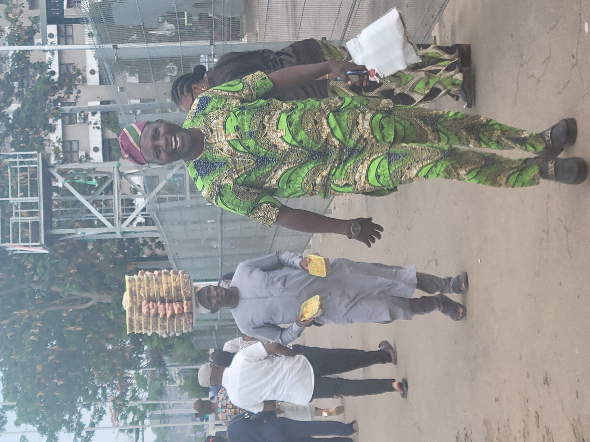 The image shows a street scene in Abuja, Nigeria. A man in a green patterned outfit and a maroon hat is smiling at the camera. Beside him, another person, partially visible, is wearing similar patterned clothing. Further away, a man in a grey outfit is carrying a stack of yellow-wrapped items on his head and holding two more in his hands. Several other people are walking in the background, dressed in various attire. The environment appears to be a busy street with some construction or metal framework visible in the background.
