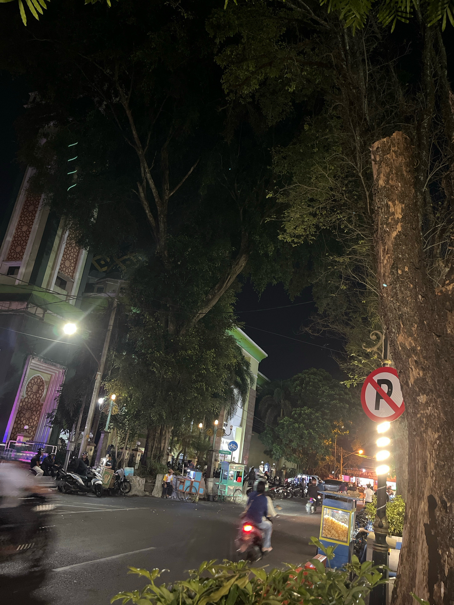 The image depicts a street scene at night in Salatiga, Indonesia. A prominent tree trunk with rough bark occupies the right side of the frame, with a "no parking" sign mounted on it. Beyond the tree, a street is visible with several motorcycles parked and moving, some with riders. In the background, a building with Islamic architectural elements, adorned with intricate geometric patterns, is illuminated. Other buildings, streetlights, and more trees are present, contributing to a vibrant, urban night atmosphere. The presence of food carts and shops suggests a commercial area. The ground appears wet, possibly from recent rain, reflecting the lights.