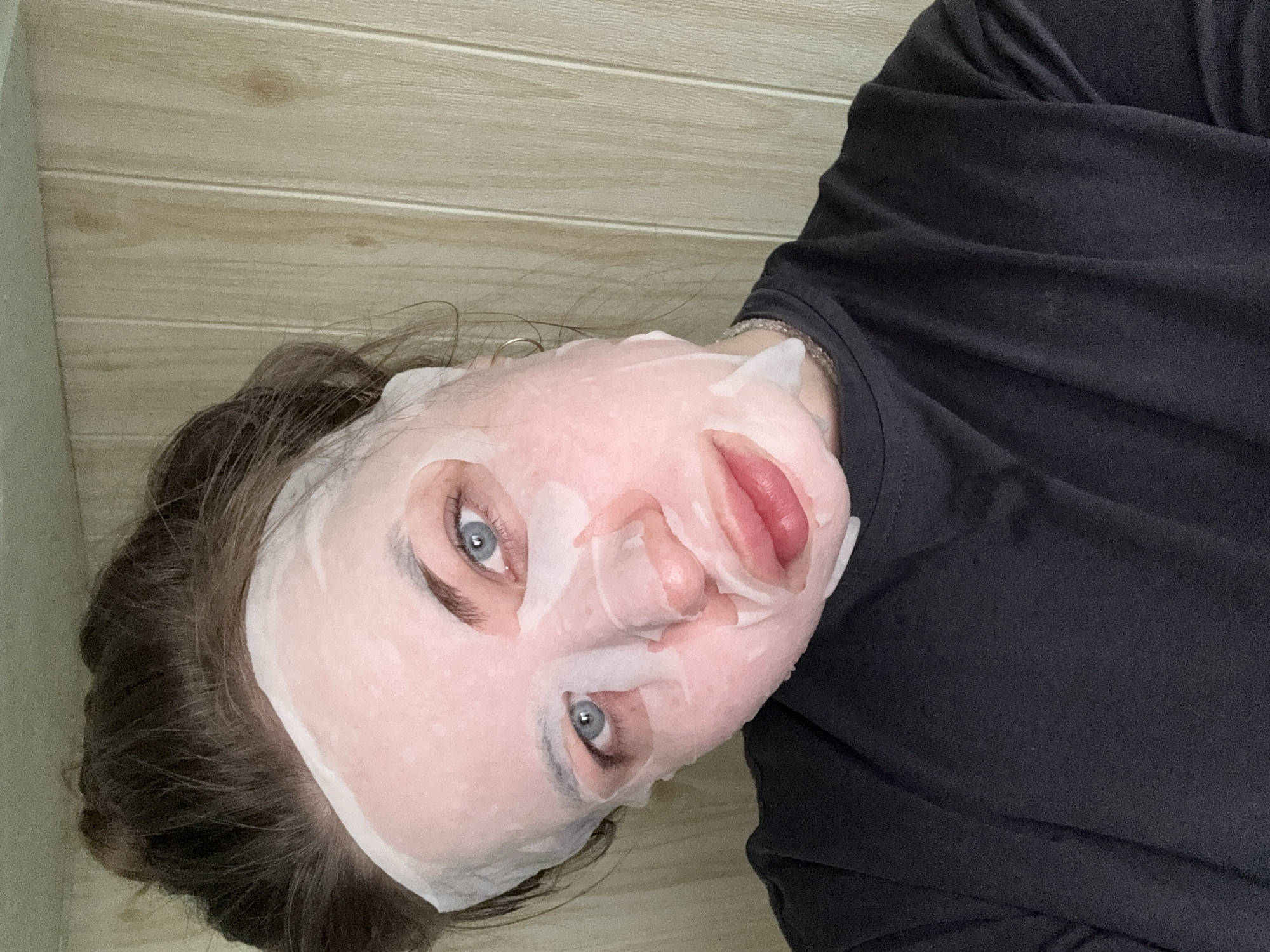 A person with a sheet face mask is photographed indoors against a wall with horizontal wood paneling. The person is wearing a dark, possibly black, shirt. The face mask is white and translucent, covering most of the face but revealing the eyes and lips. The eyes are blue. The hair around the mask is dark brown. The location is Chaikovsky, Russia.