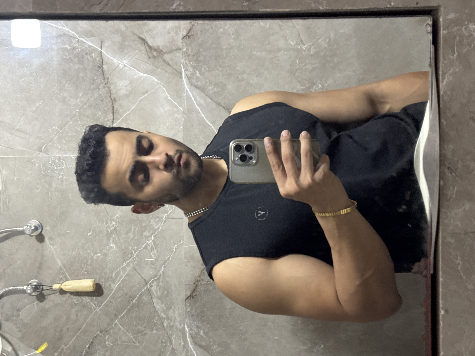 A man is reflected in a bathroom mirror. He is wearing a black tank top and a gold bracelet on his left wrist. He is holding a cell phone in his right hand, presumably taking a selfie. The bathroom features gray, marble-patterned walls. There is a towel rack and other bathroom fixtures visible on the wall behind him. The location is unknown, and there is no indication in the picture that it is in Kolkata, India.