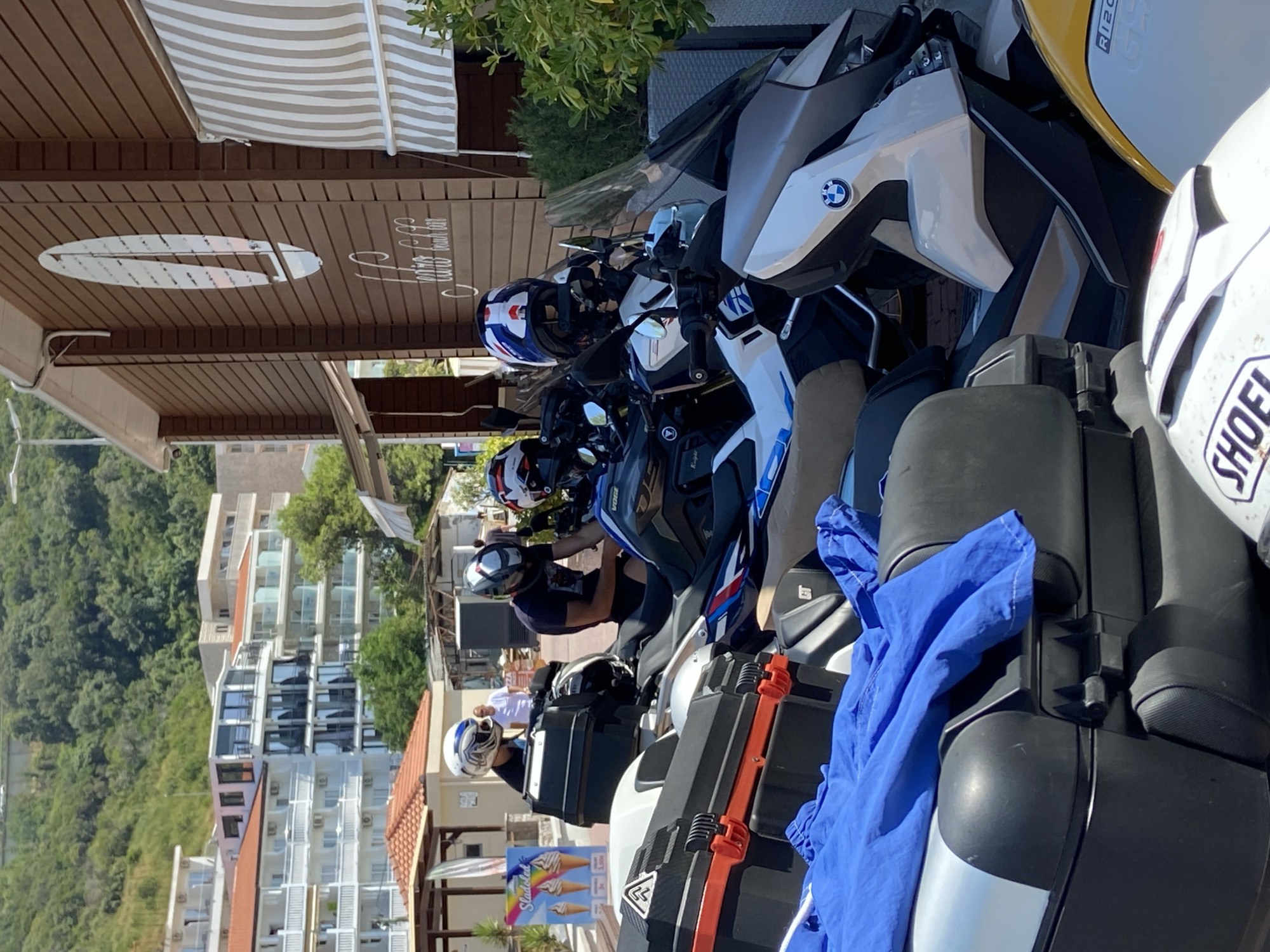 In Prijevor, Montenegro, a group of individuals, possibly tourists, are gathered near parked motorcycles. Three motorcycles are visible, two with prominent black storage cases on the rear, one of which is covered by a blue cloth. Two individuals are wearing helmets, one with a white, blue, and red design and another with a white and blue pattern. The motorcycles appear to be adventure or touring models. In the background, a multi-story building with white balconies and orange-tiled roofs is visible, suggesting a coastal or resort area. The facade of a brown building with striped awnings and a circular emblem is also present, potentially a cafe or restaurant. A sign with images of ice cream cones and prices is partially visible, indicating a nearby establishment. The scene is set on a sunny day, with lush green trees and vegetation adding to the environment.