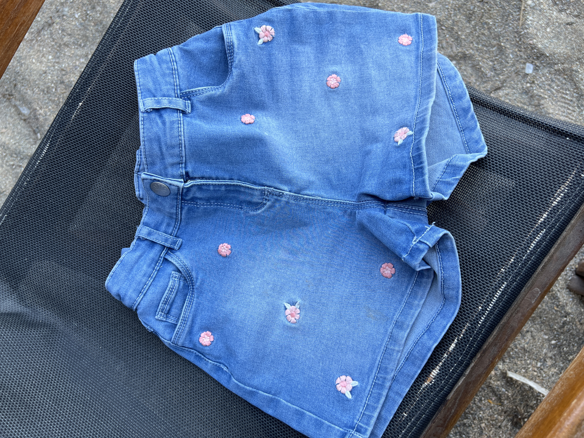 A pair of blue denim shorts with embroidered pink flowers is displayed on a dark mesh surface, likely part of outdoor seating. The shorts are laid flat, showing the front pockets and waistband with a button closure. The background consists of sandy ground and parts of wooden furniture, suggesting an outdoor setting. The location is identified as Eretria Municipal Unit, Greece.