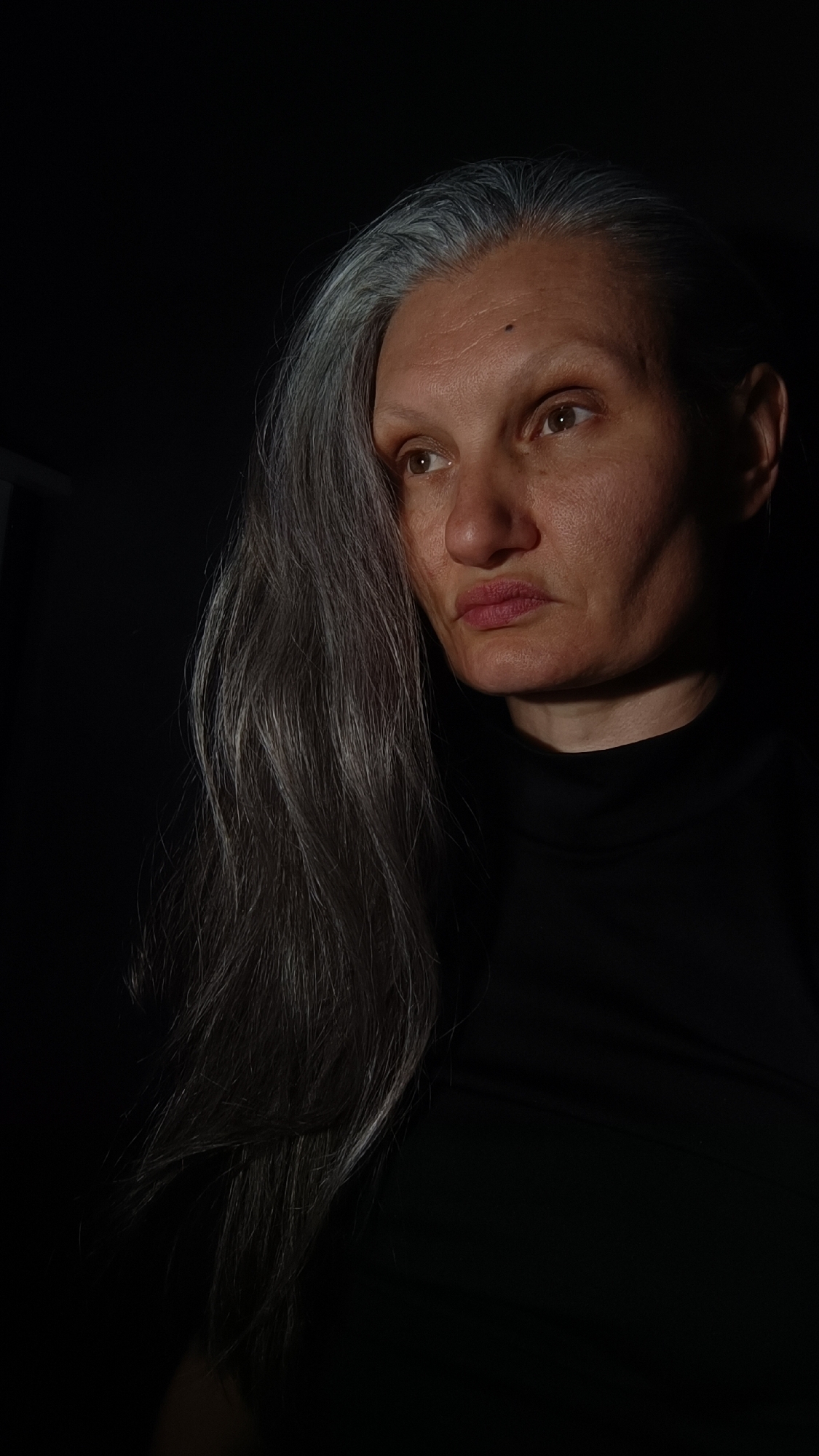 A close-up portrait depicts a woman with long, gray hair. She wears a black turtleneck shirt. Her facial features are clearly visible, with particular attention to her skin texture and the lines around her eyes and mouth. The lighting is dramatic, with strong shadows that create a sense of depth and emphasize the contours of her face. The background is dark and indistinct. There is no indication that the photo was taken in Lermontov, Russia or any other place.
