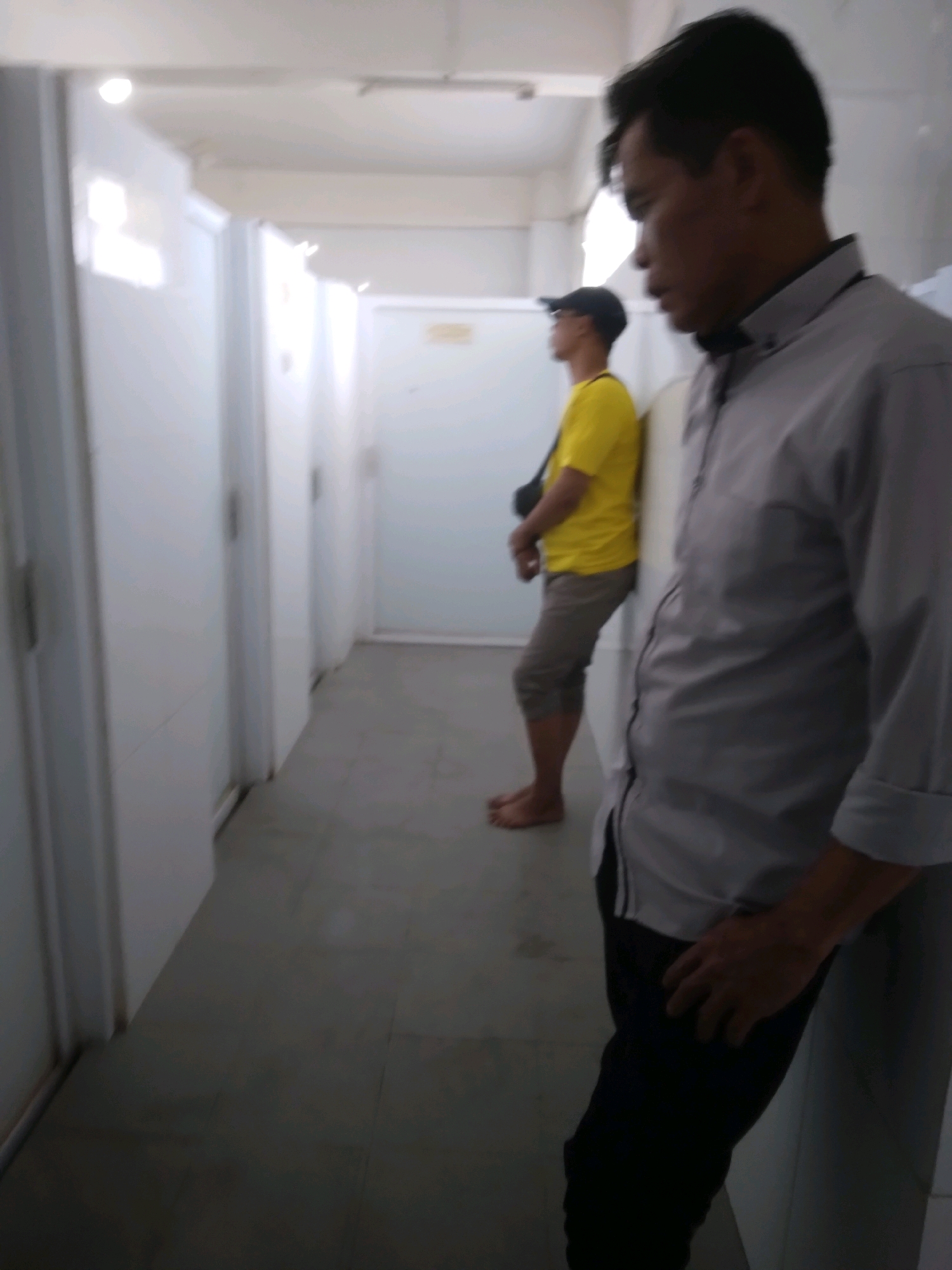 The image depicts an indoor hallway with several white doors lining the left side. A man in the foreground, wearing a gray collared shirt and dark pants, leans against a wall on the right. His body is partially turned away from the camera, with his right hand resting on his leg. In the background, further down the hallway, a second man stands near a white door, leaning against it. He is wearing a yellow t-shirt, tan shorts, and a dark baseball cap. His left arm is visible, and he is barefoot. The hallway floor is tiled in a gray, checkered pattern. The lighting appears to be from overhead fluorescent lights, casting a shadow across the floor. The scene is consistent with a public restroom facility or a row of rooms in a communal building. The location is identified as Sokaraja, Indonesia, suggesting a public facility within that area.