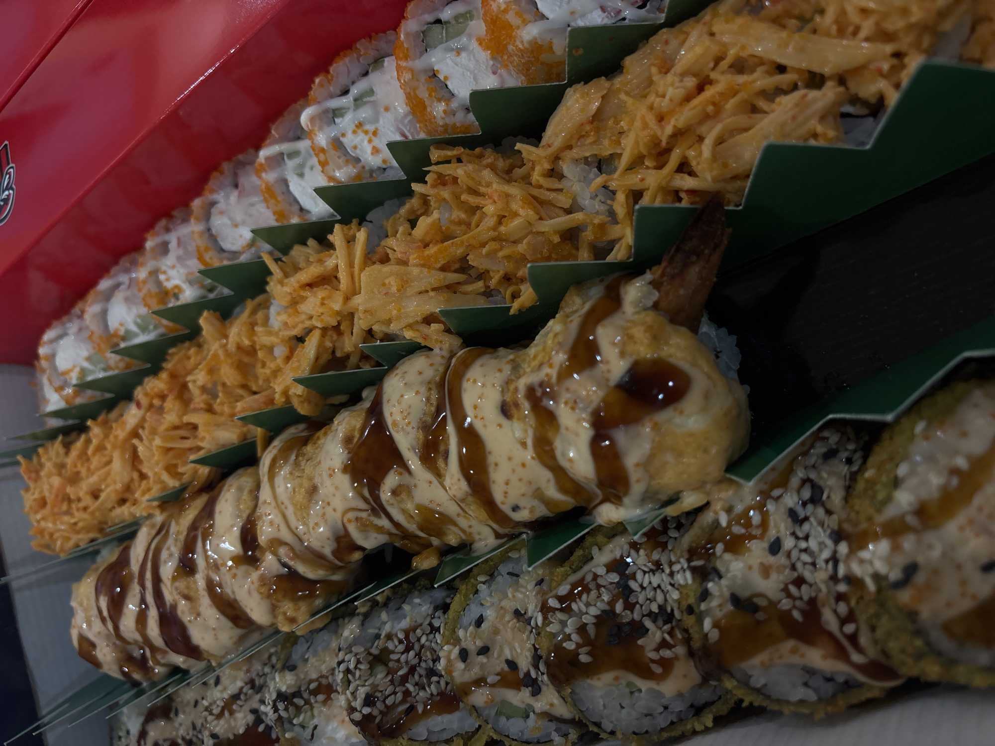 This close-up, high-angle shot captures a vibrant assortment of sushi rolls meticulously arranged within a red takeout box, separated by green zig-zag dividers. The image displays at least four distinct types of sushi, presented for a meal or consumption.
In the background, rows of California-style rolls are visible, featuring white rice exteriors, a light creamy sauce, and numerous bright orange specks, likely tobiko or fish roe. Adjacent to these are rolls generously topped with an abundance of thin, crispy, light orange-yellow strands, which could be fried surimi, tempura flakes, or crispy fried onions, providing a textural contrast.
The mid-ground highlights a prominent, elongated sushi roll, possibly a specialty item, coated in a thick, creamy sauce and elegantly drizzled with a darker, glossy sauce. A visible shrimp tail suggests it might be a tempura shrimp roll. In the foreground, further rows of sushi rolls are visible, characterized by a golden, crispy exterior, indicating they are likely tempura-fried. These rolls are garnished with a mix of white and black sesame seeds and finished with the same creamy and dark sauce drizzles seen on the elongated roll.
The sushi is neatly compartmentalized by the green dividers, emphasizing an organized and appealing presentation. A partially visible, stylized red and white logo is present on the red box in the upper left corner, but its specific text or design is not clearly discernible. The overall scene focuses on the variety, detailed preparation, and appealing presentation of the sushi rolls.